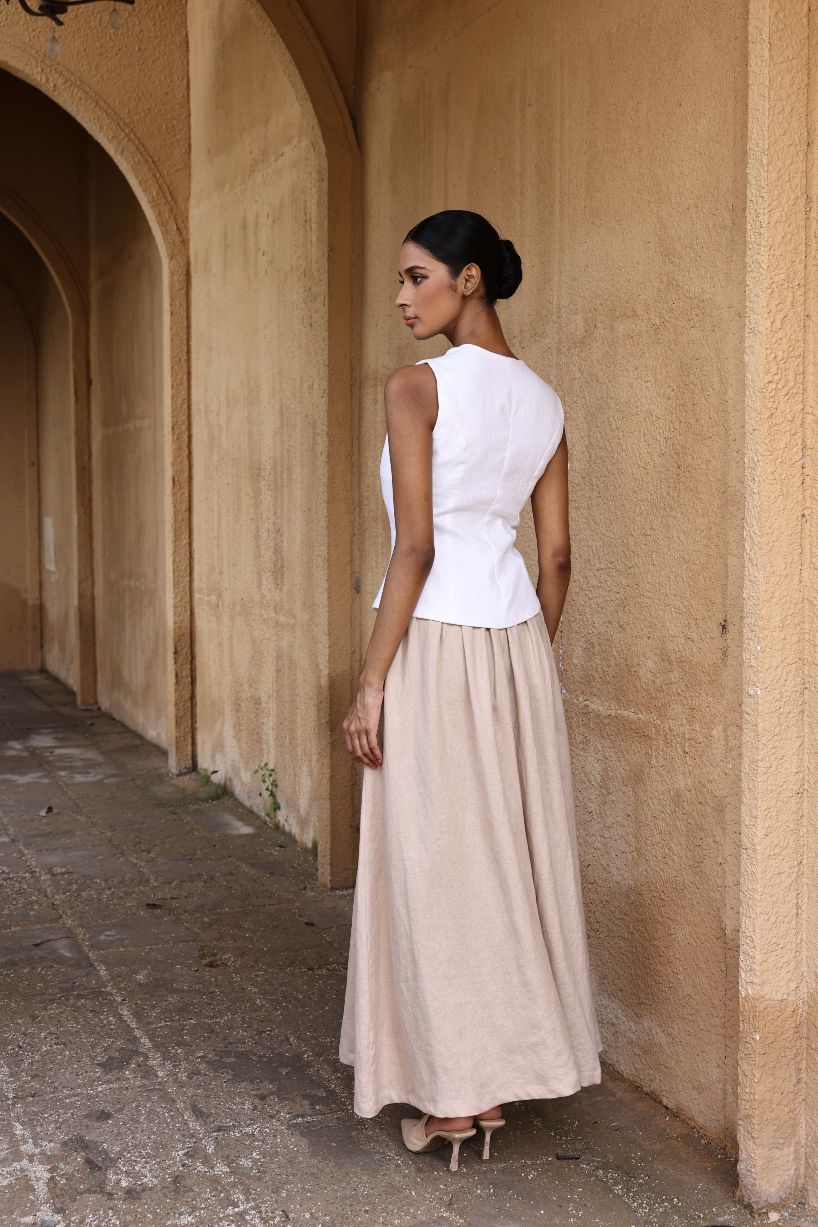 Woman in a white top and beige skirt standing against a beige wall.