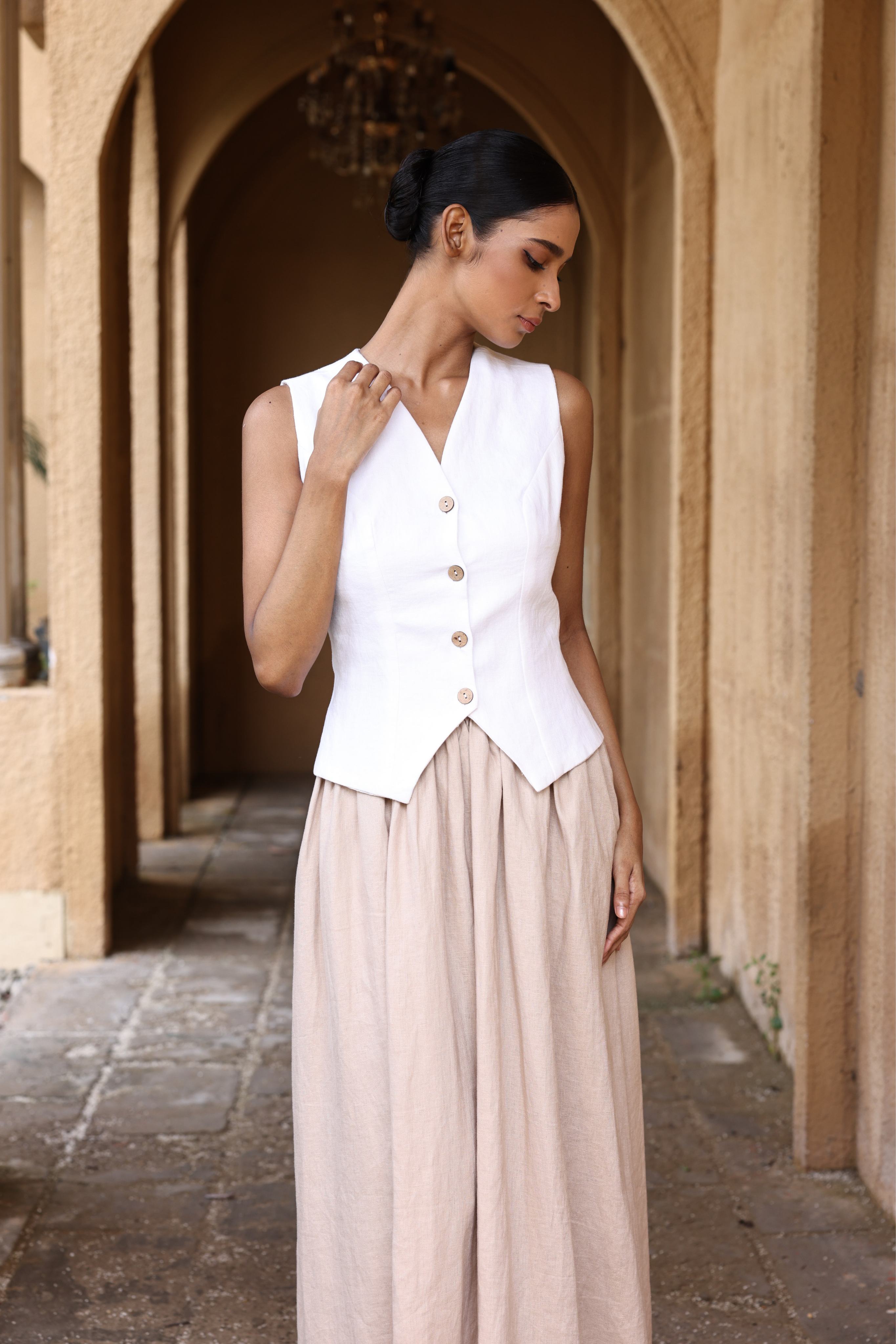 Woman wearing a white vest and beige skirt standing in an arched stone doorway.