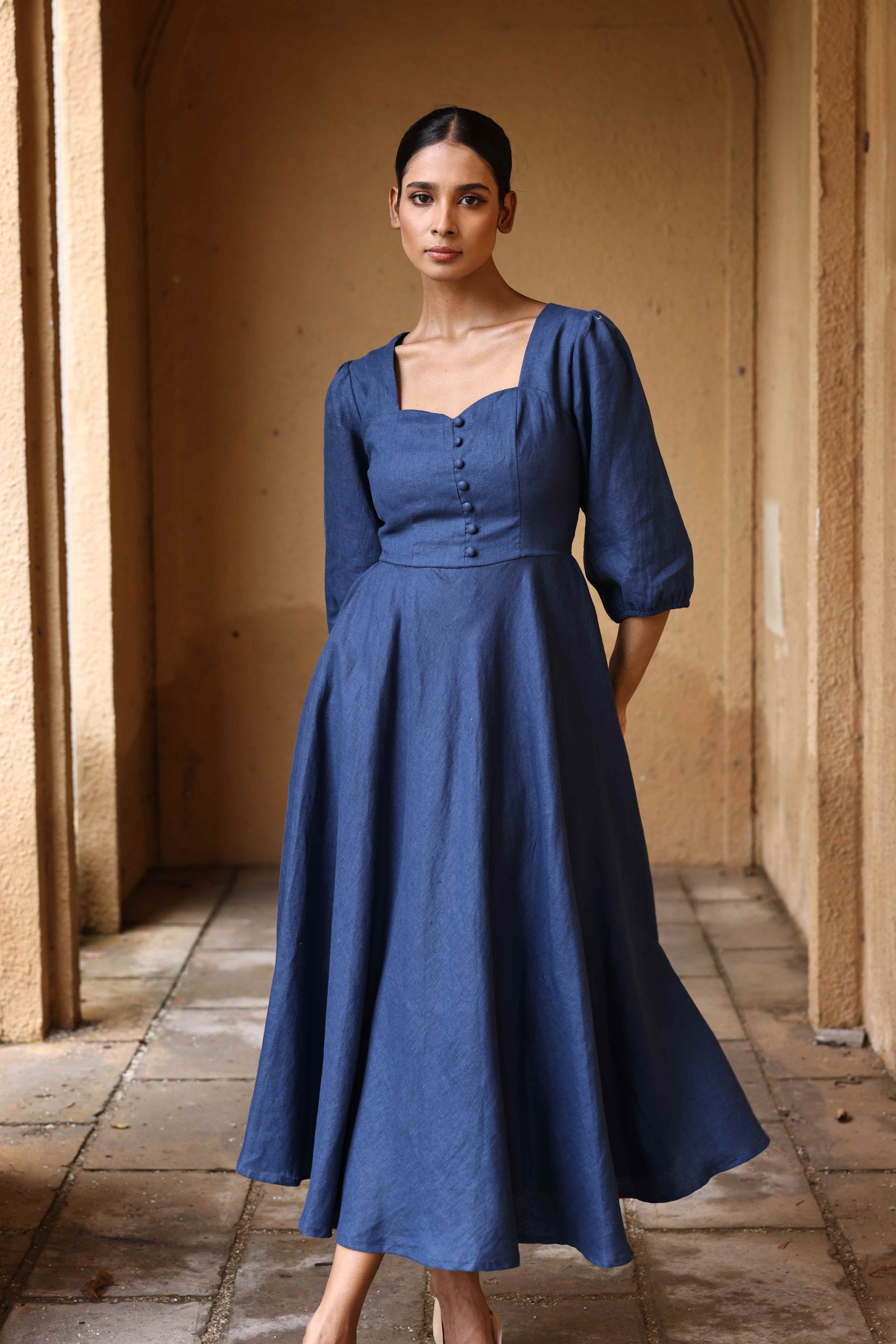 A woman wearing a blue dress with a high neckline and puffed sleeves stands in a hallway.