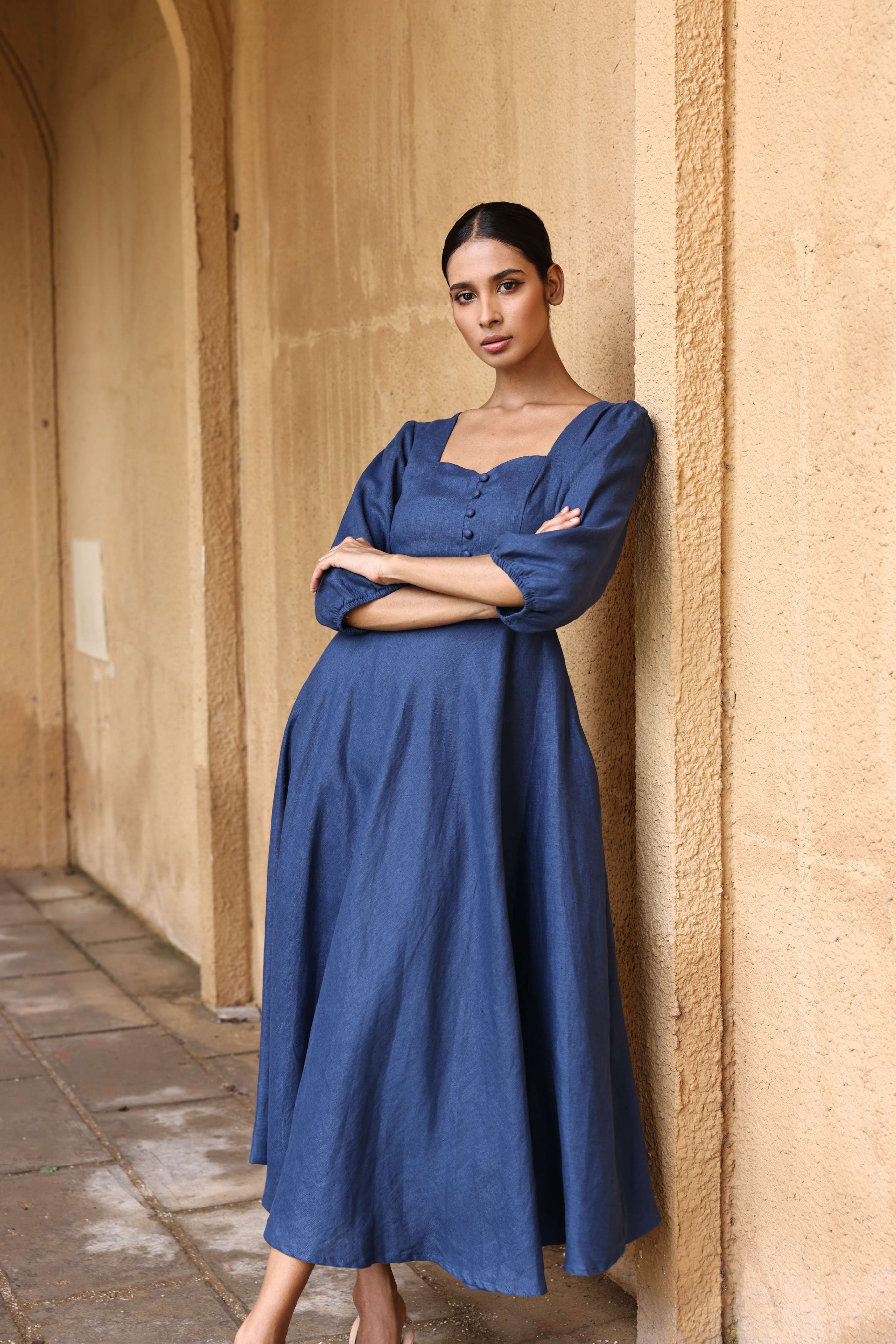A woman in a blue dress stands with her arms crossed, leaning against a beige wall.