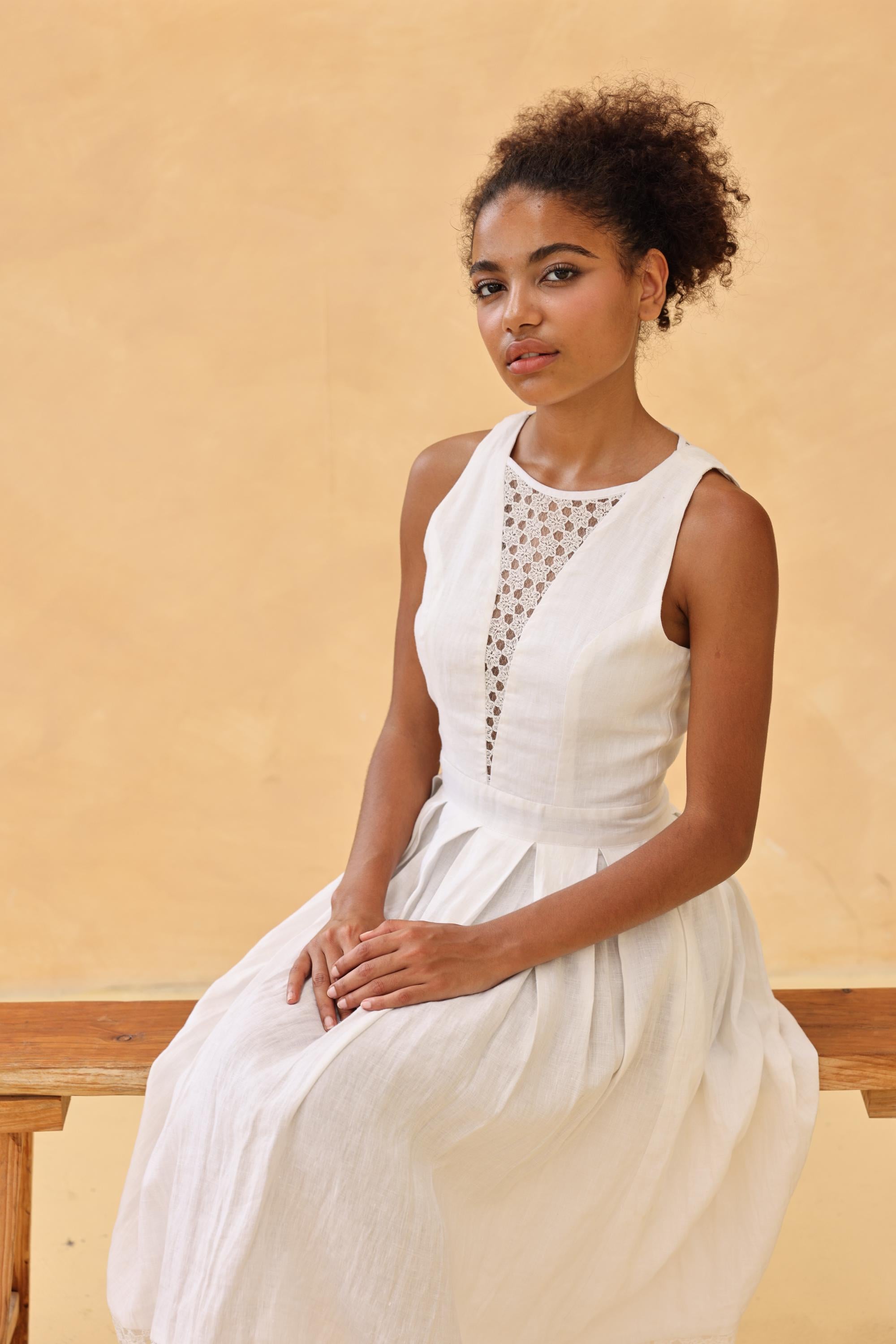 Woman in a white dress sitting on a wooden bench against a beige wall