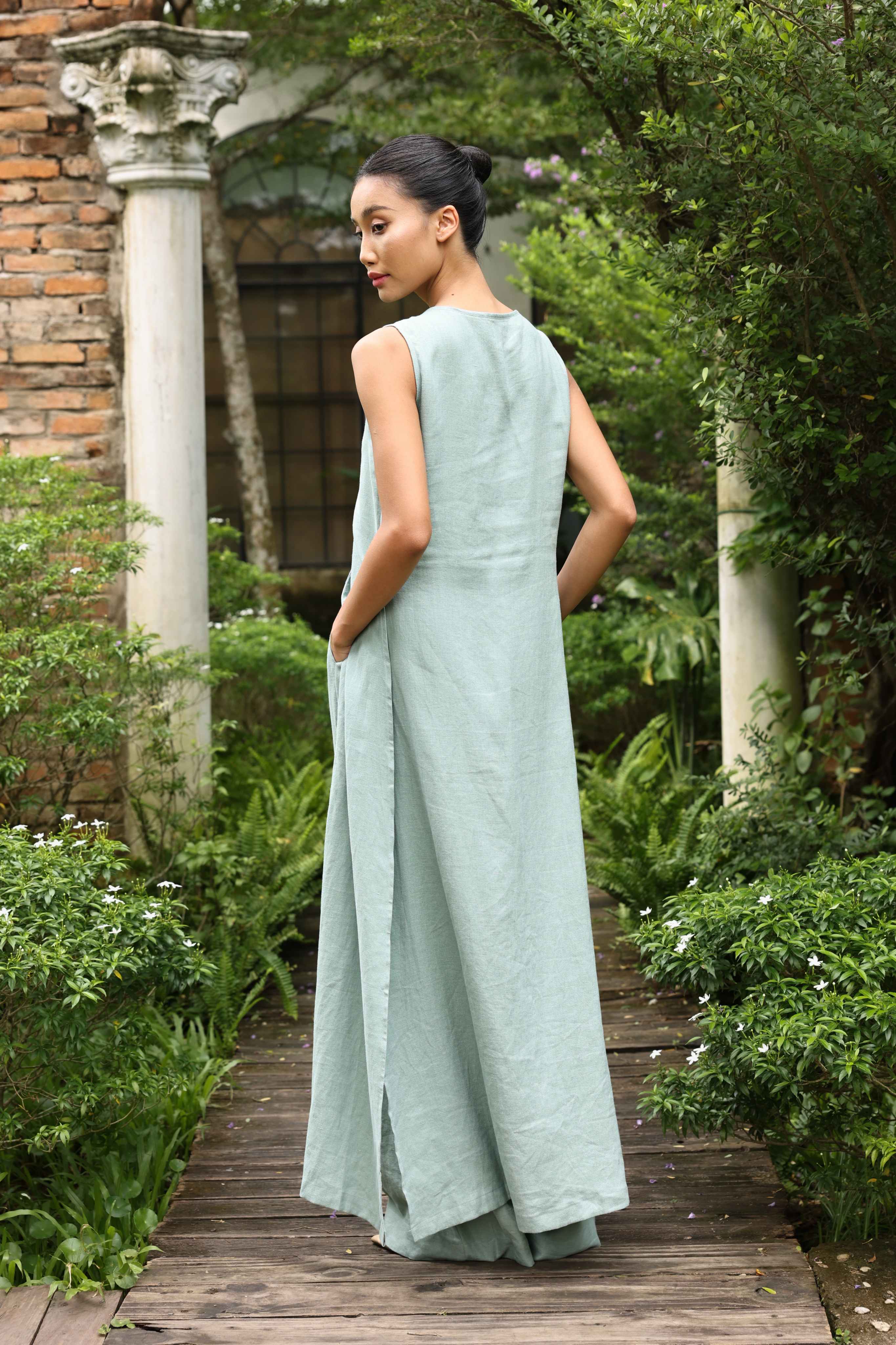 Woman in a light green tunic standing on a wooden path in a garden setting.