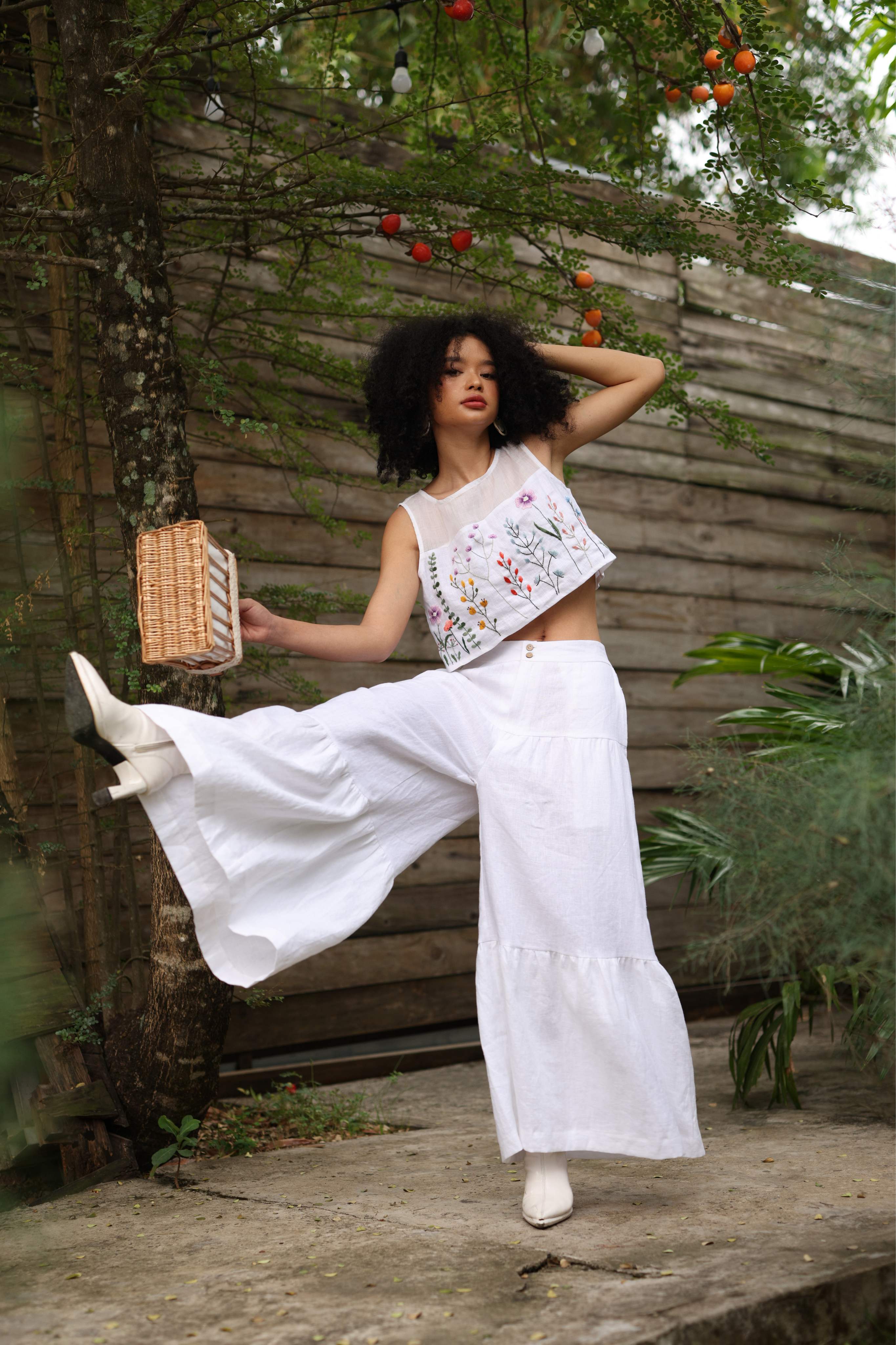 Woman in a white outfit with floral embroidery standing outdoors near a wooden fence and fruit tree.