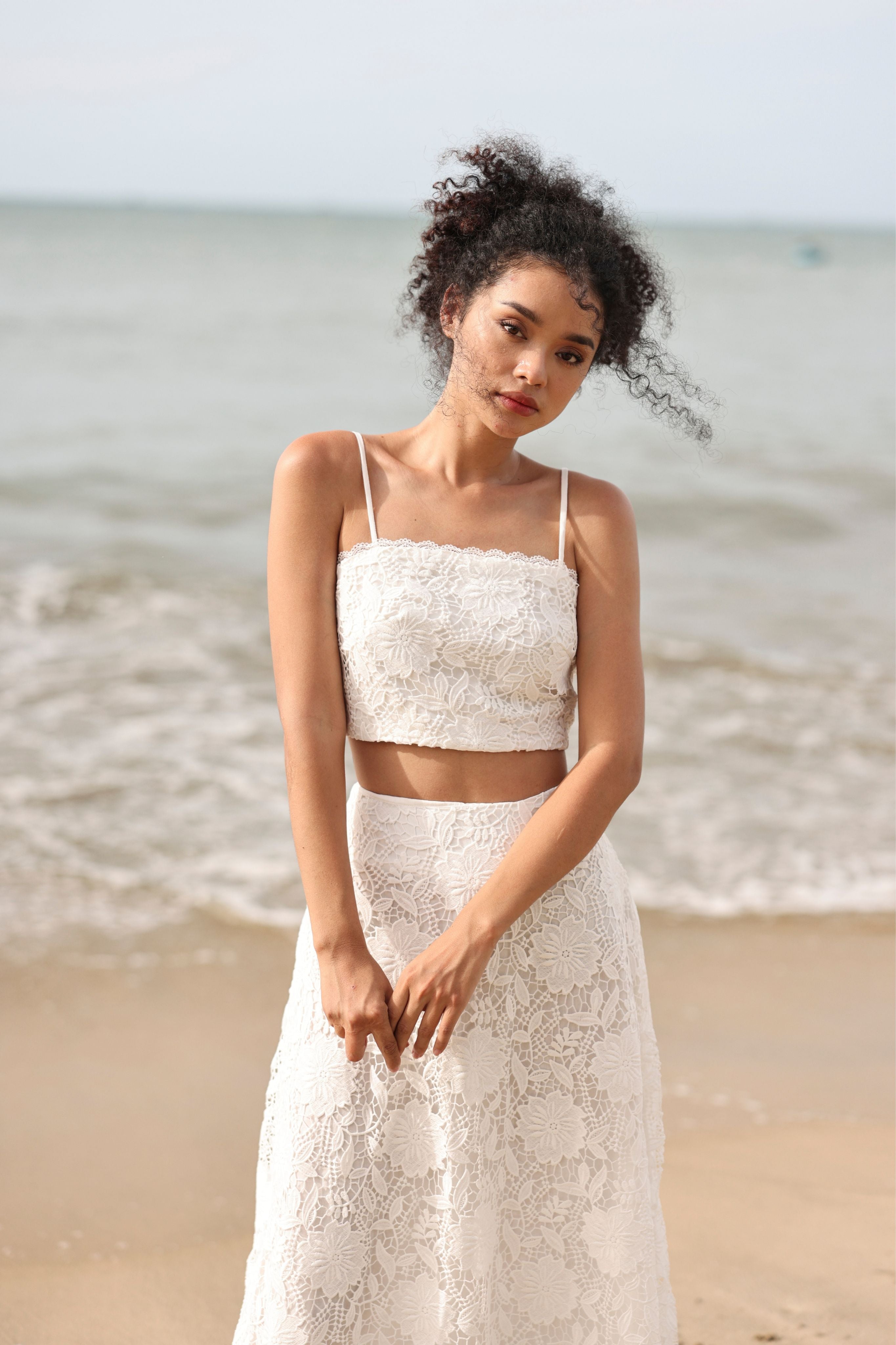 Woman in a white lace top and skirt standing on a beach with ocean waves in the background