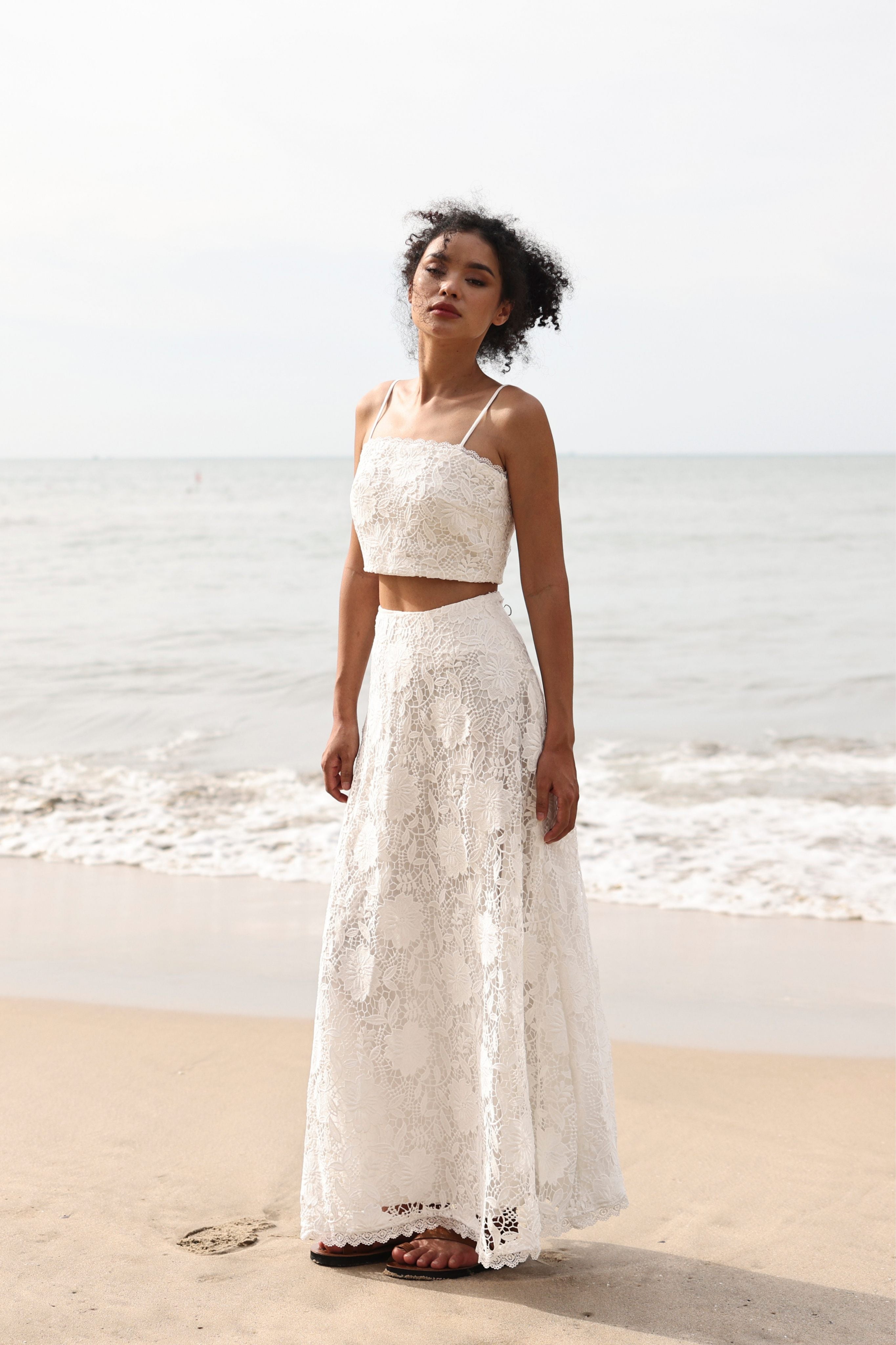 Woman in a white lace top and skirt standing on a beach with ocean in the background