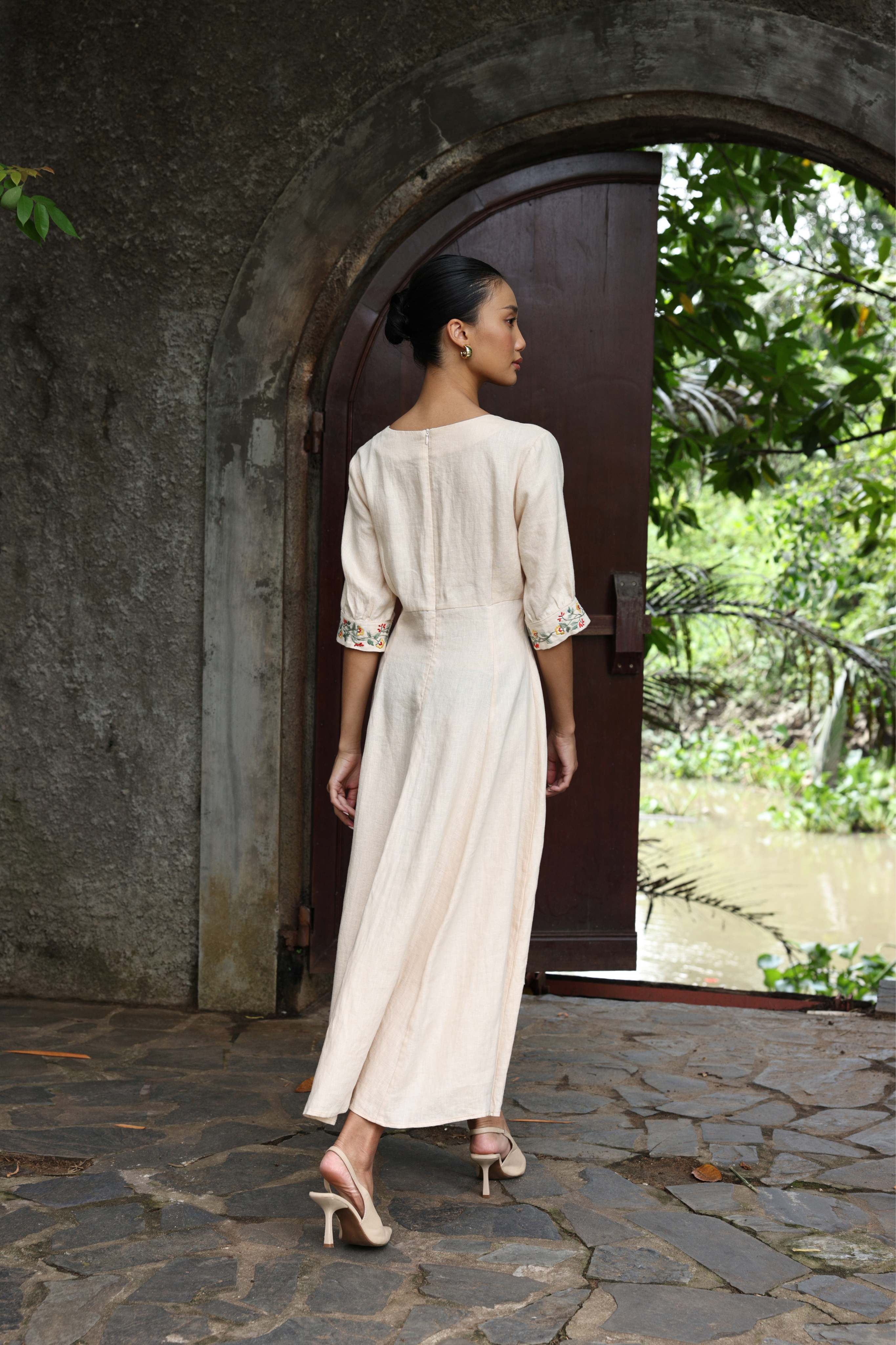 Woman in a long, light-colored dress standing in front of a stone archway with greenery around.