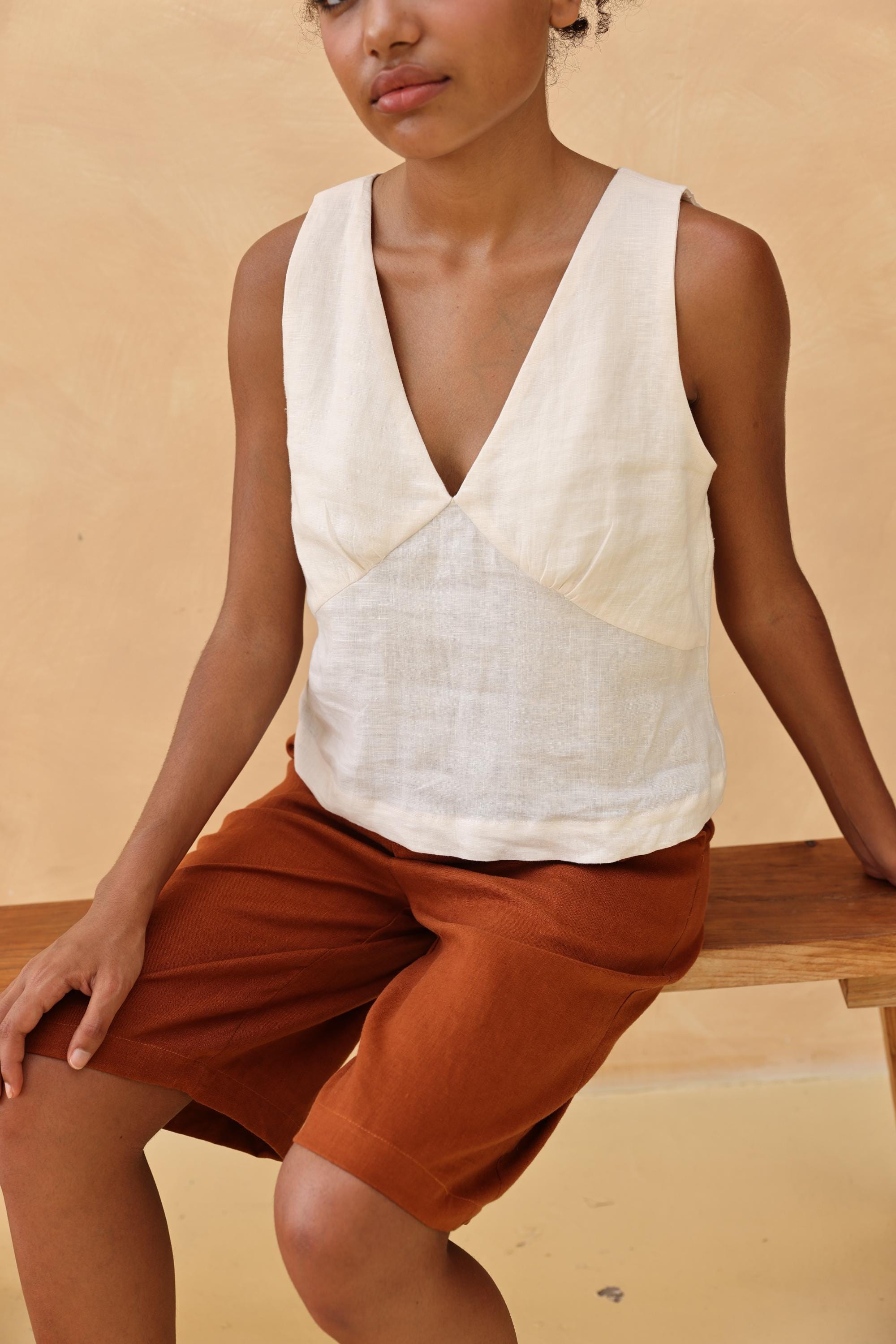 Woman wearing a sleeveless top and shorts sitting on a wooden bench against a beige background