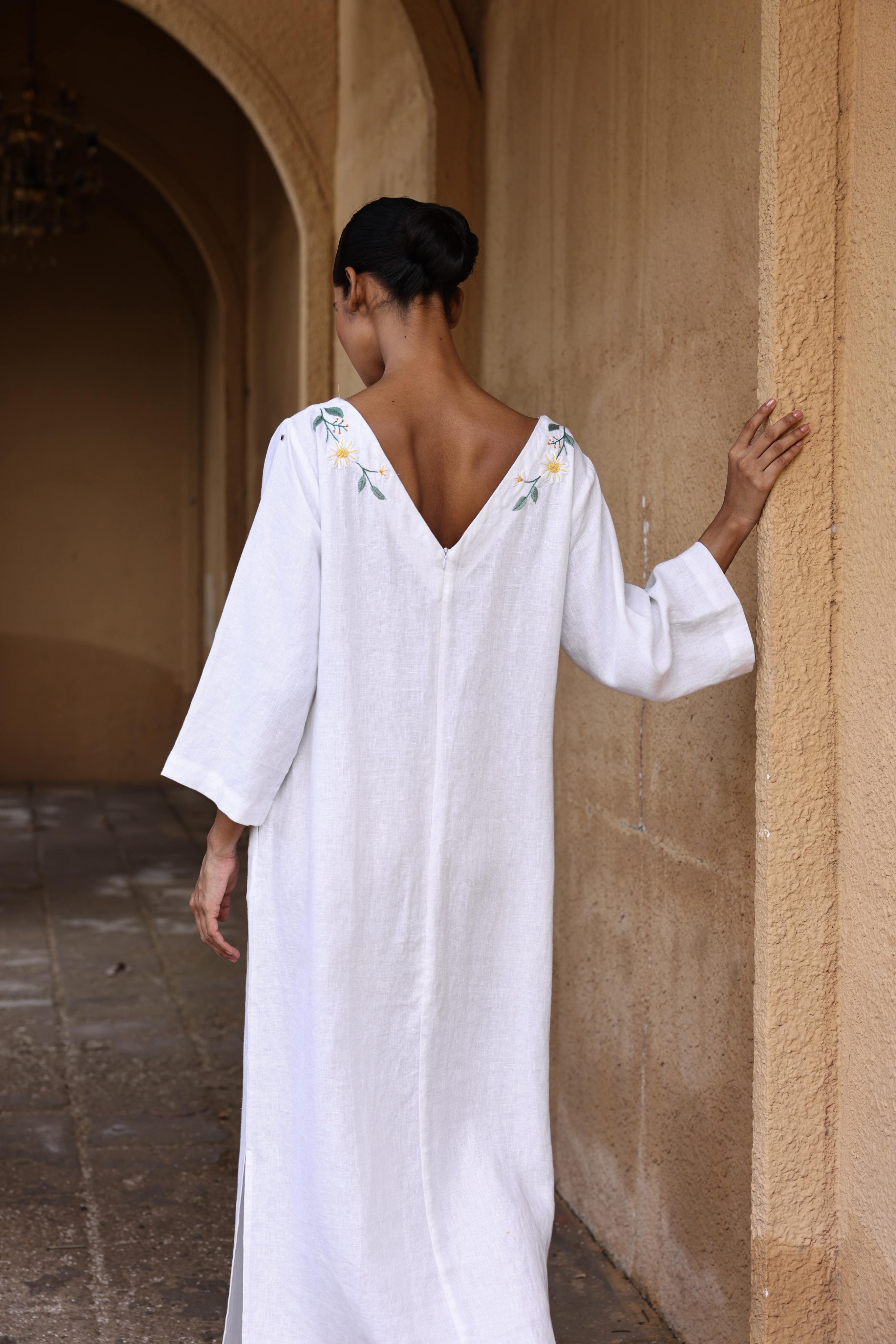Woman in a white dress with floral embroidery standing against a beige wall.