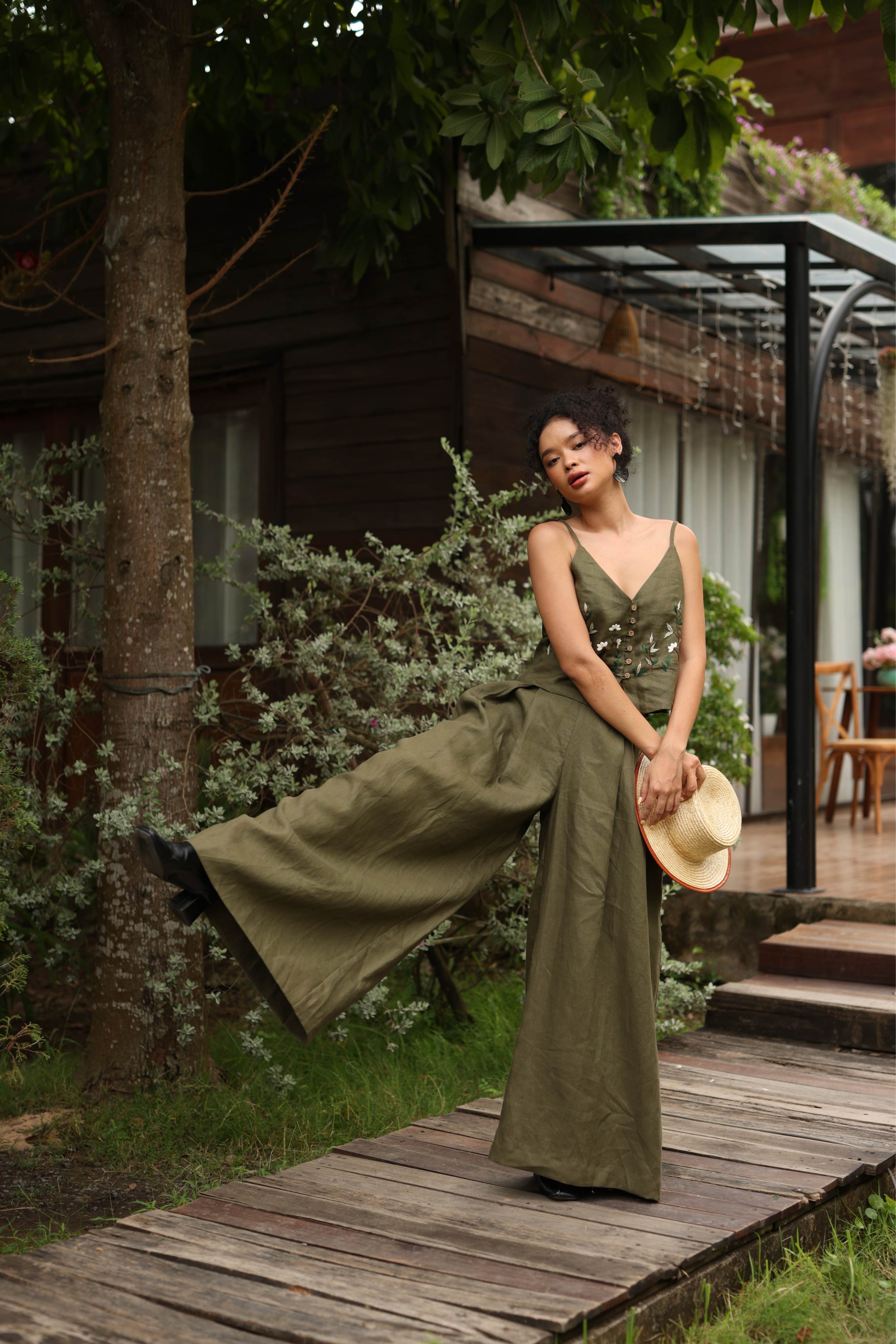 Woman in a green outfit standing on a wooden path with a building and trees in the background