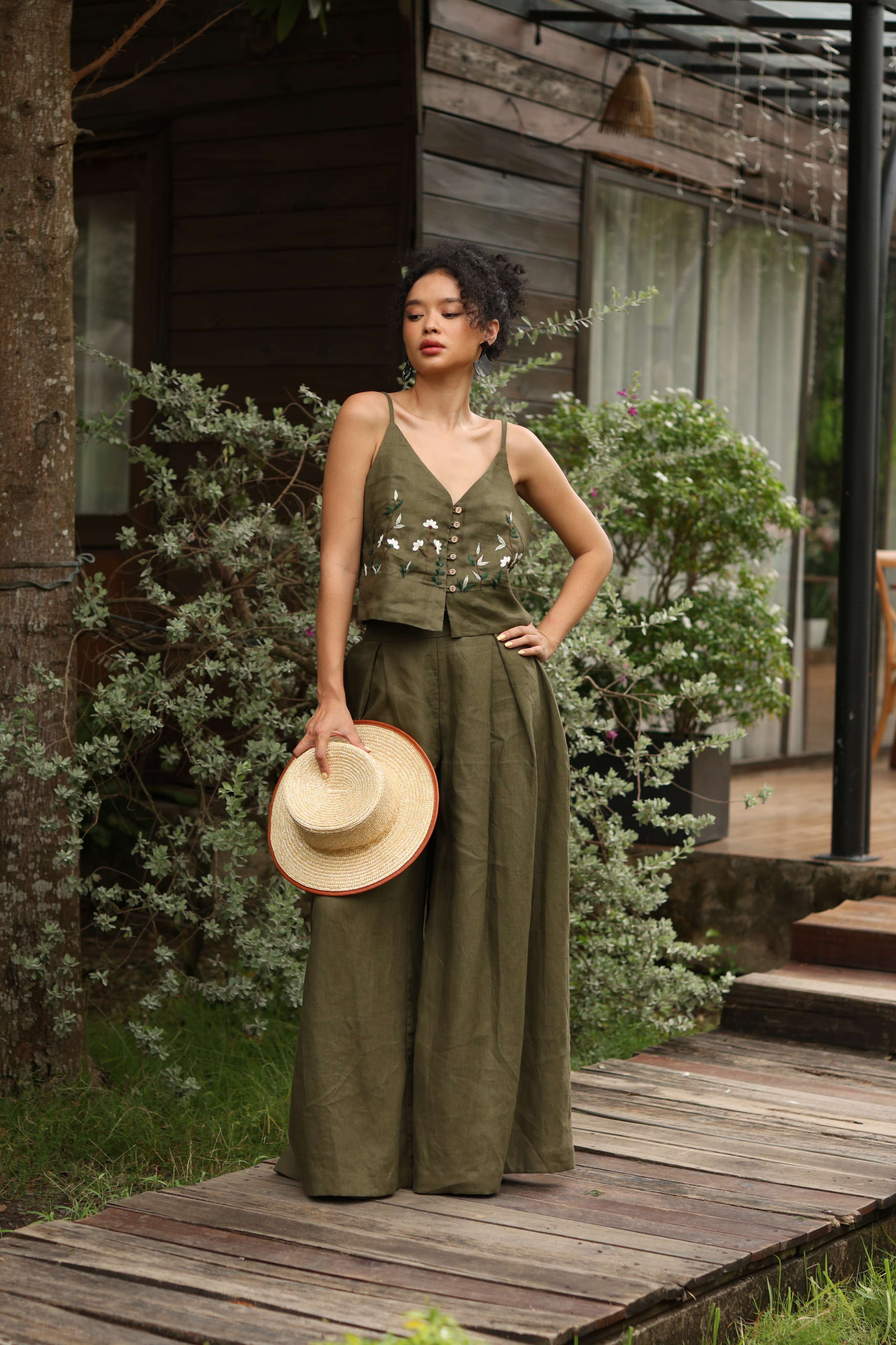 Woman in an olive green outfit holding a hat on a wooden path.