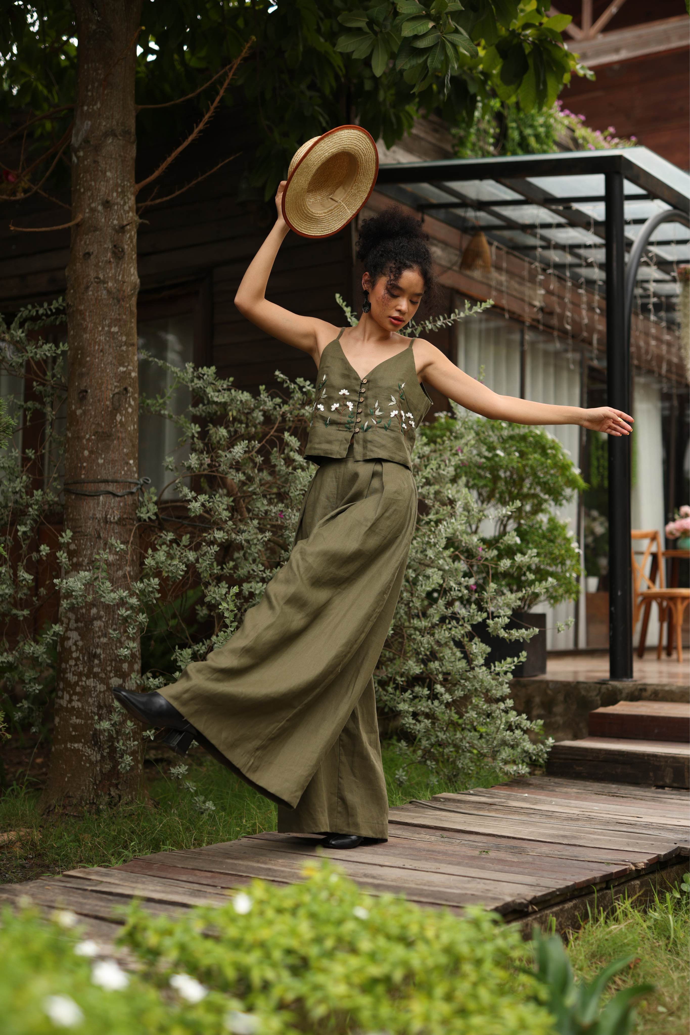 Woman in a green outfit and hat dancing outdoors on a wooden path with greenery and a building in the background.