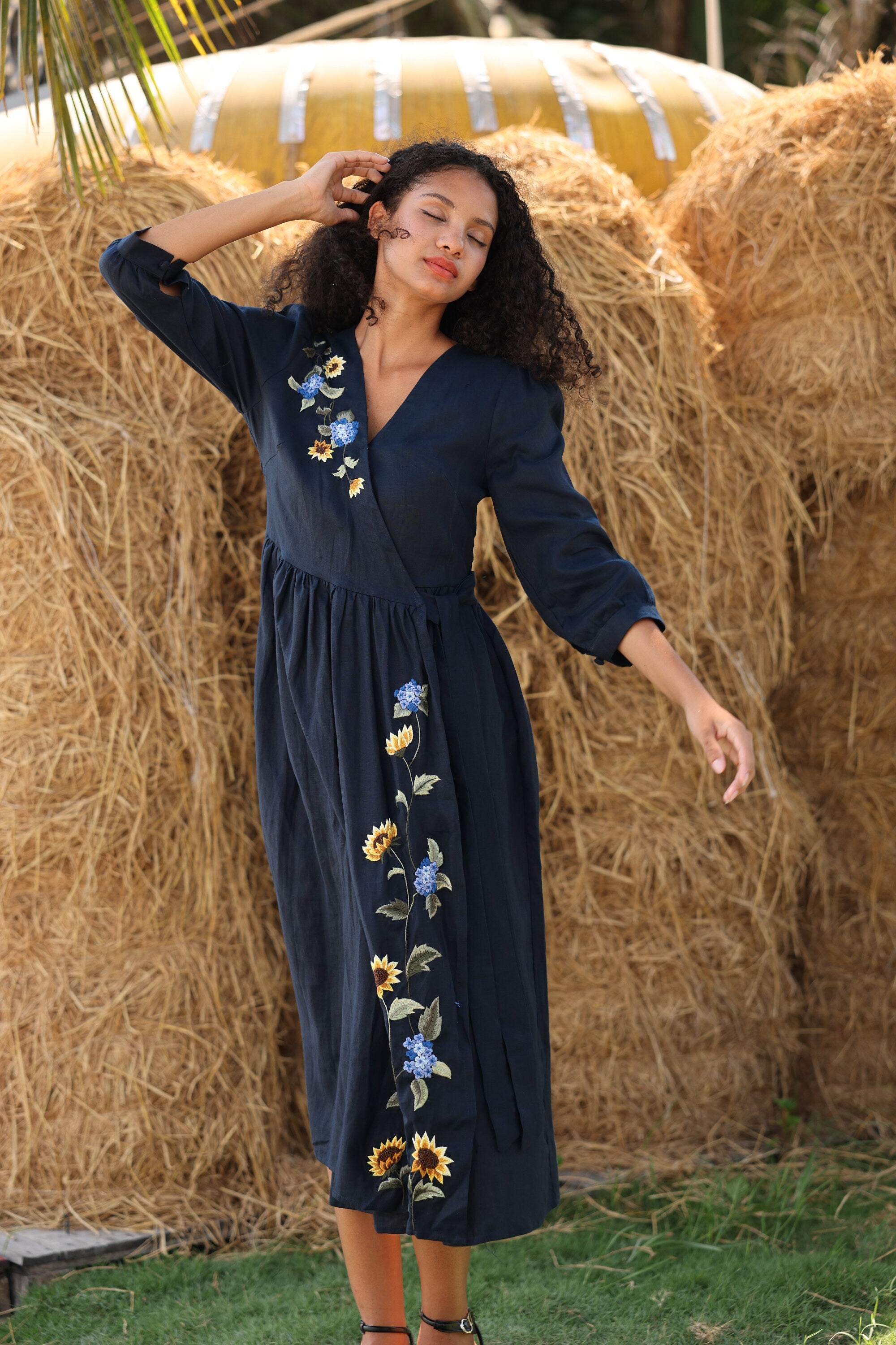 Woman in a navy dress with floral embroidery standing in front of hay bales.