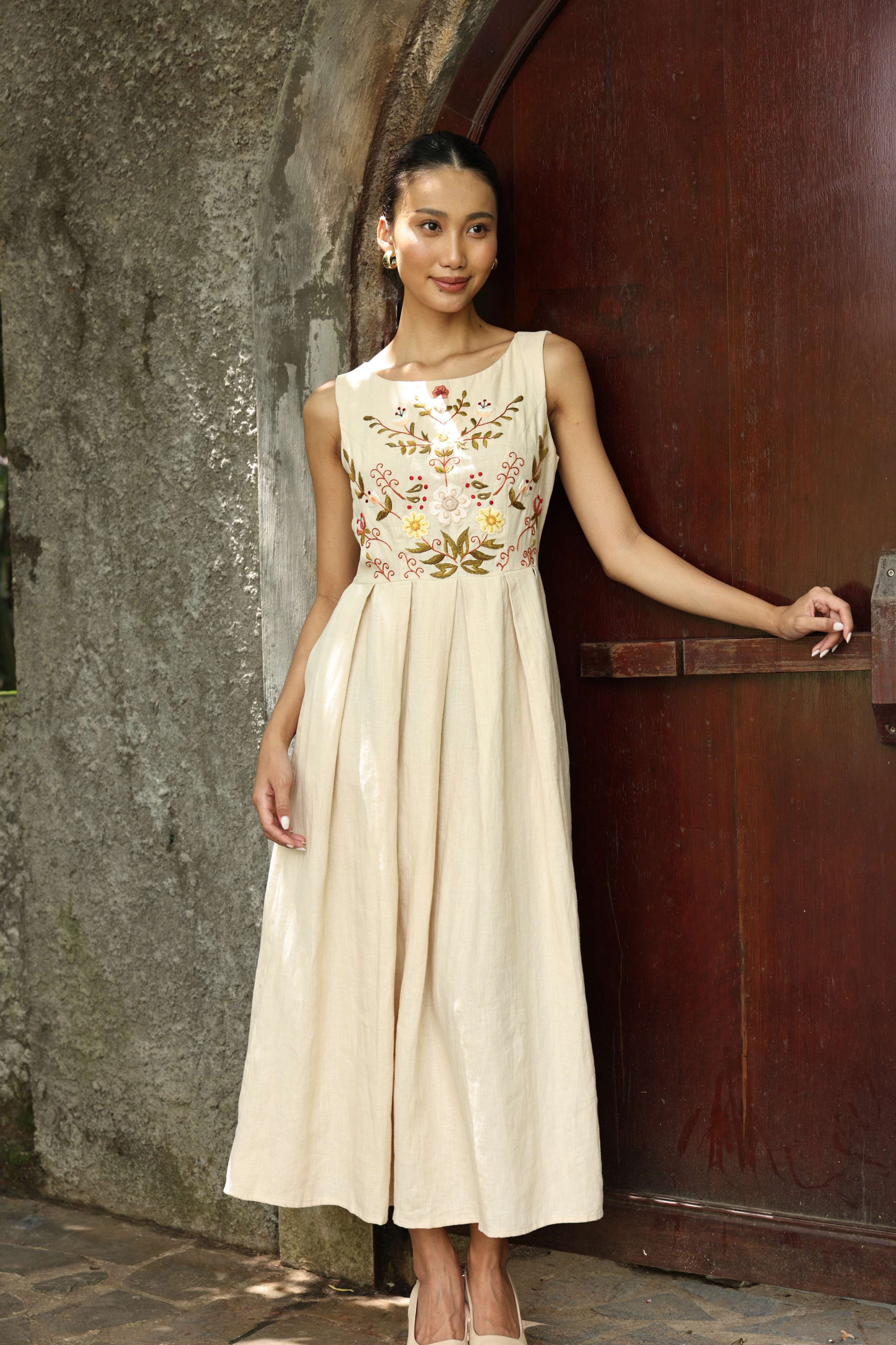 Woman in a dress with floral embroidery standing against a textured wall.