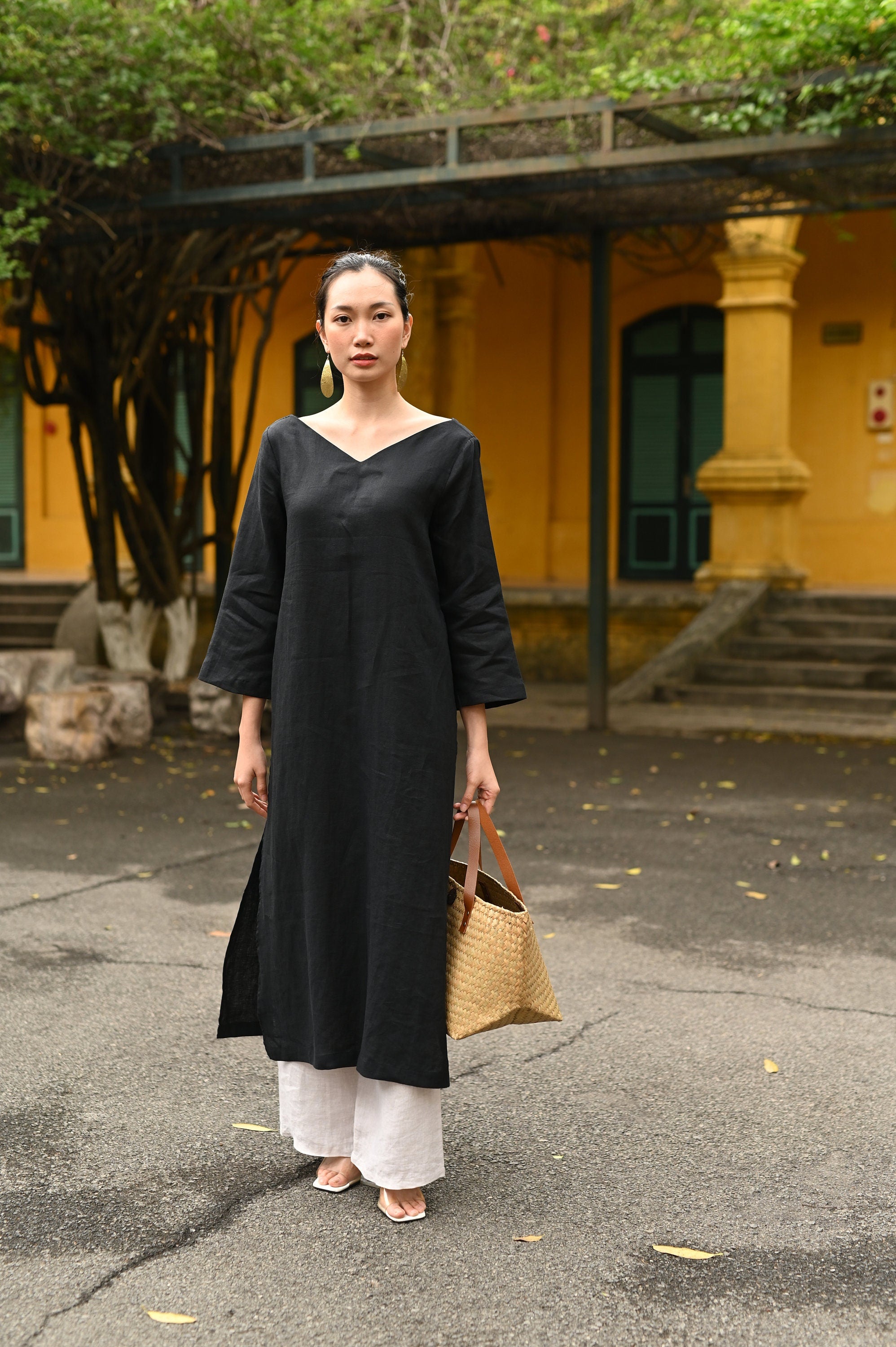 Woman in a black dress holding a woven bag outdoors with a yellow building in the background