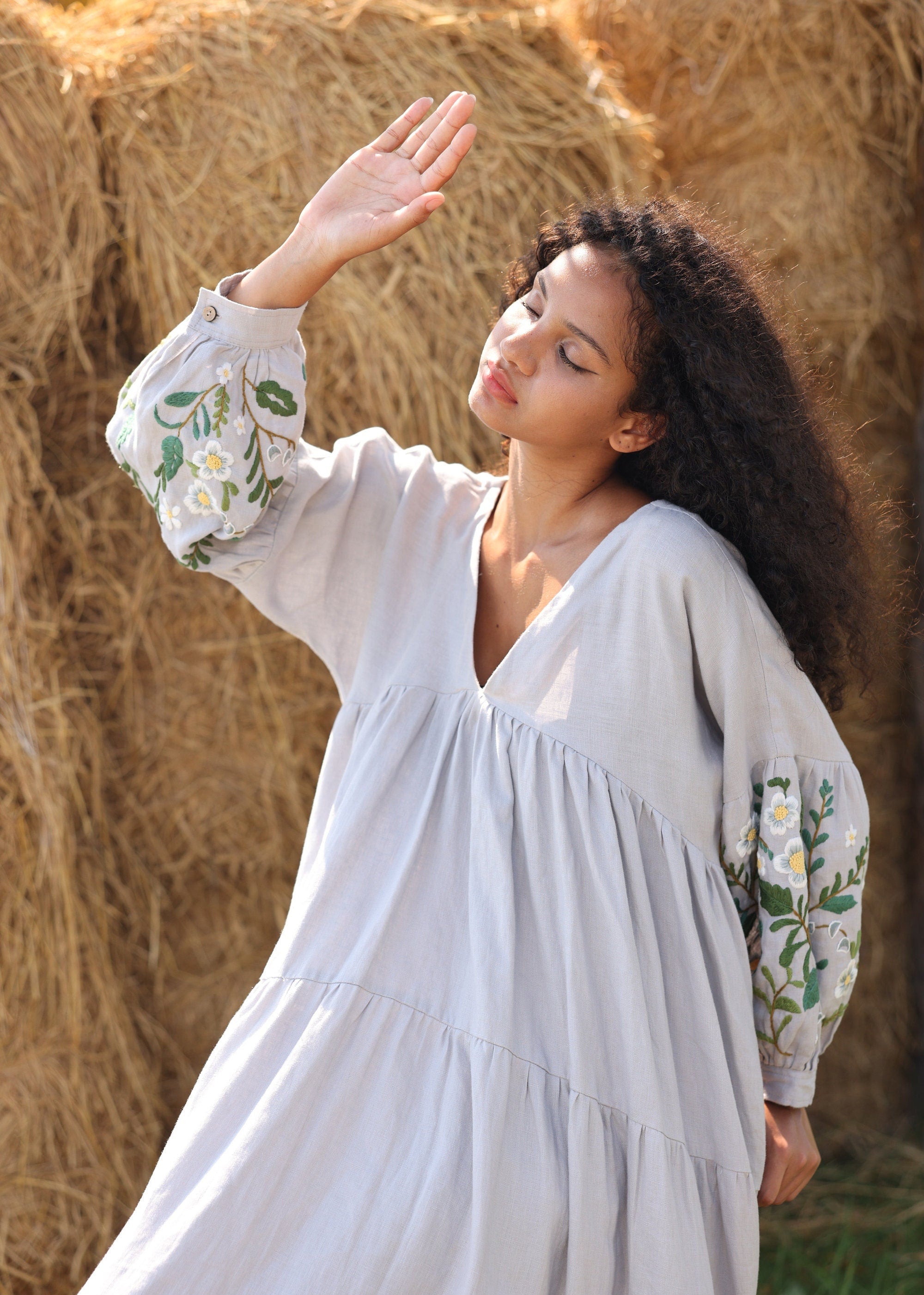 Woman in a dress with floral embroidery standing in front of hay bales.