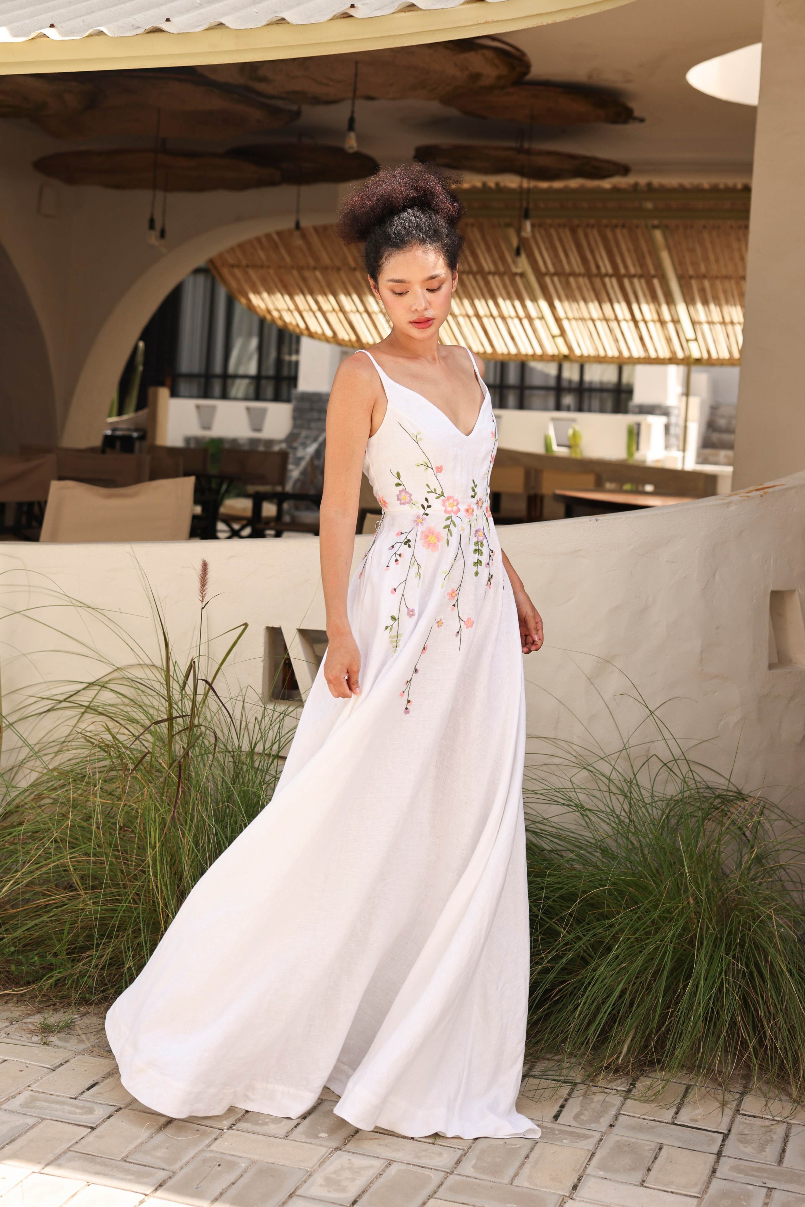 Woman in a white floral dress standing outdoors with plants and a building in the background