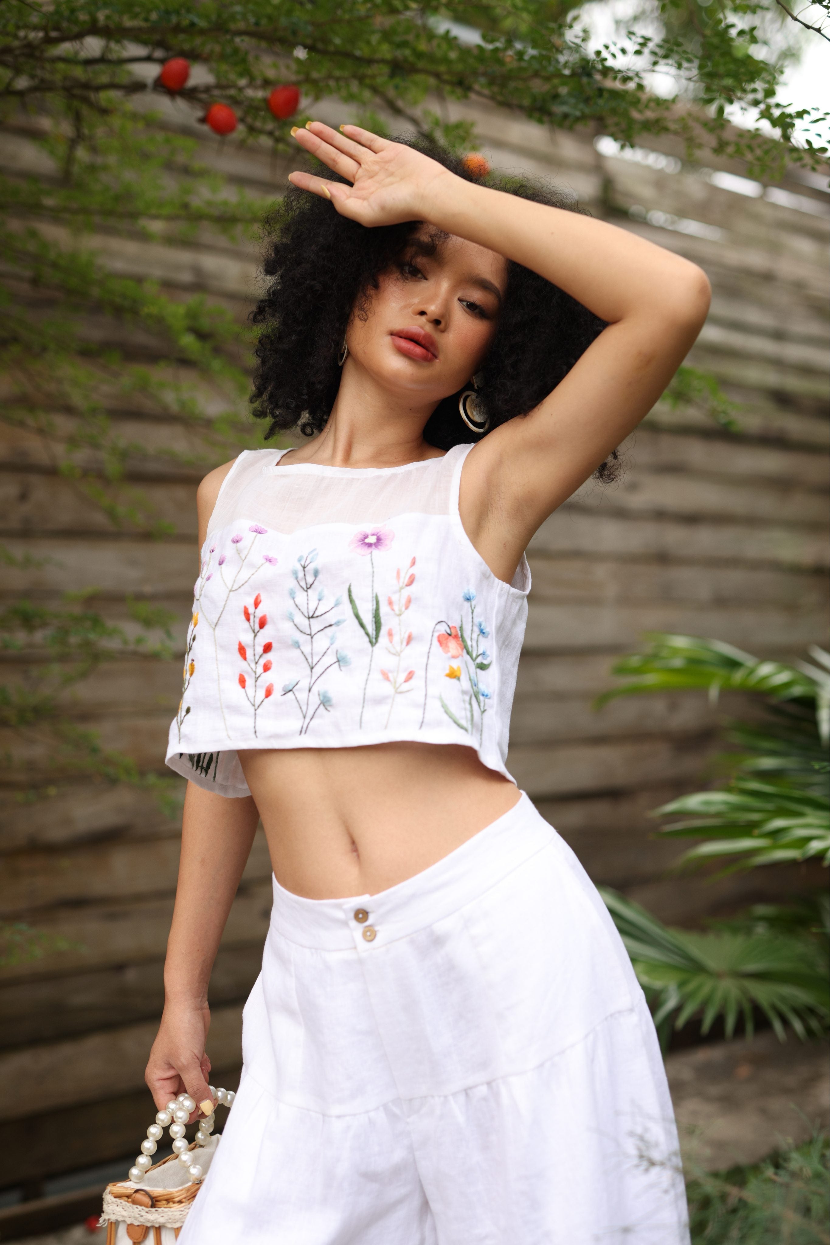 Woman wearing a white embroidered top and pants outdoors with greenery.