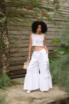 Woman in a sleeveless top and white pants standing outdoors with a wooden wall and plants in the background