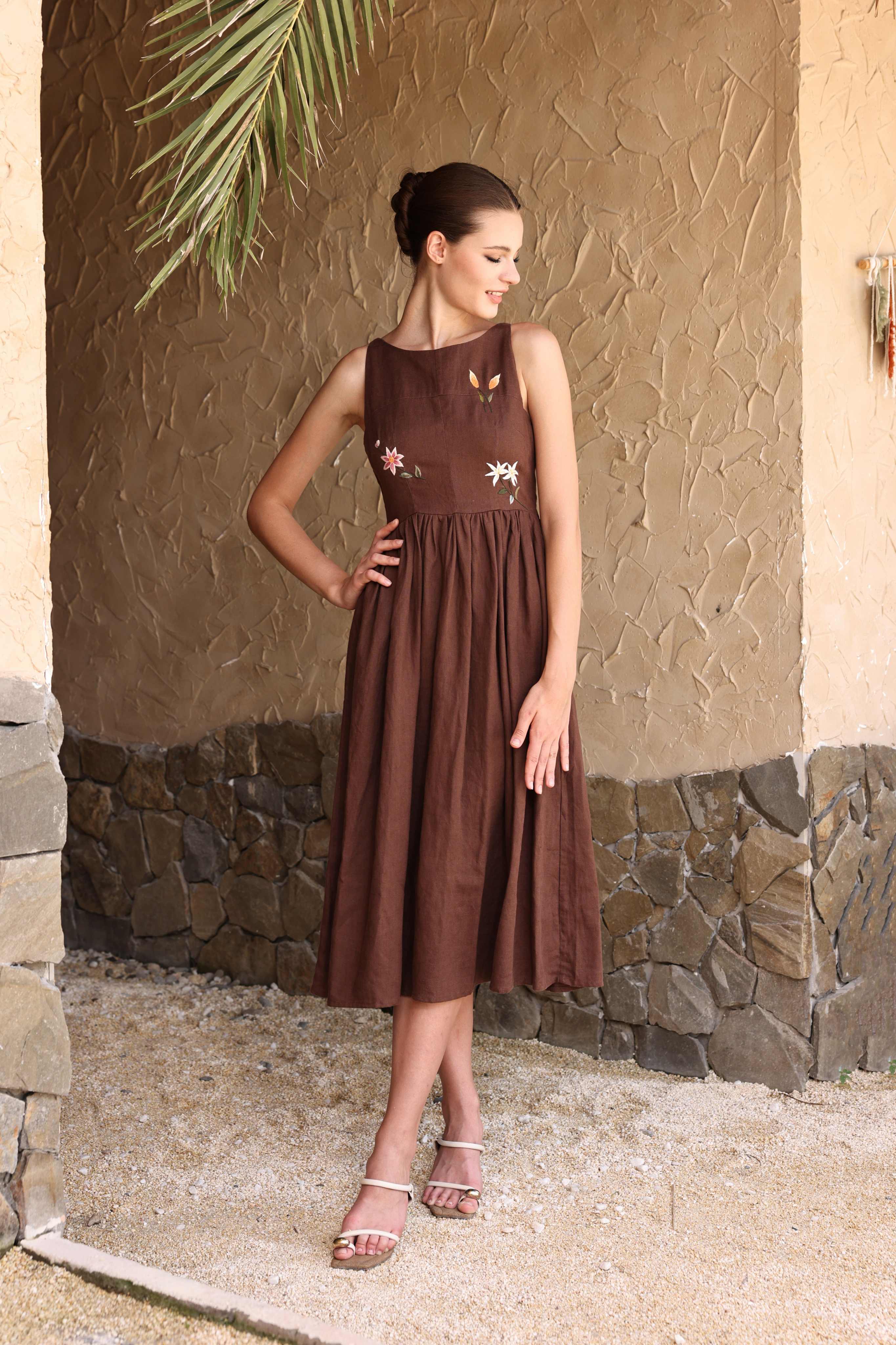 Woman wearing a brown dress with floral patterns against a stone wall.