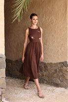 Woman wearing a brown dress with floral embellishments against a stone wall.