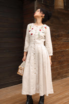 Woman wearing a beige dress with floral embroidery against a wooden wall.