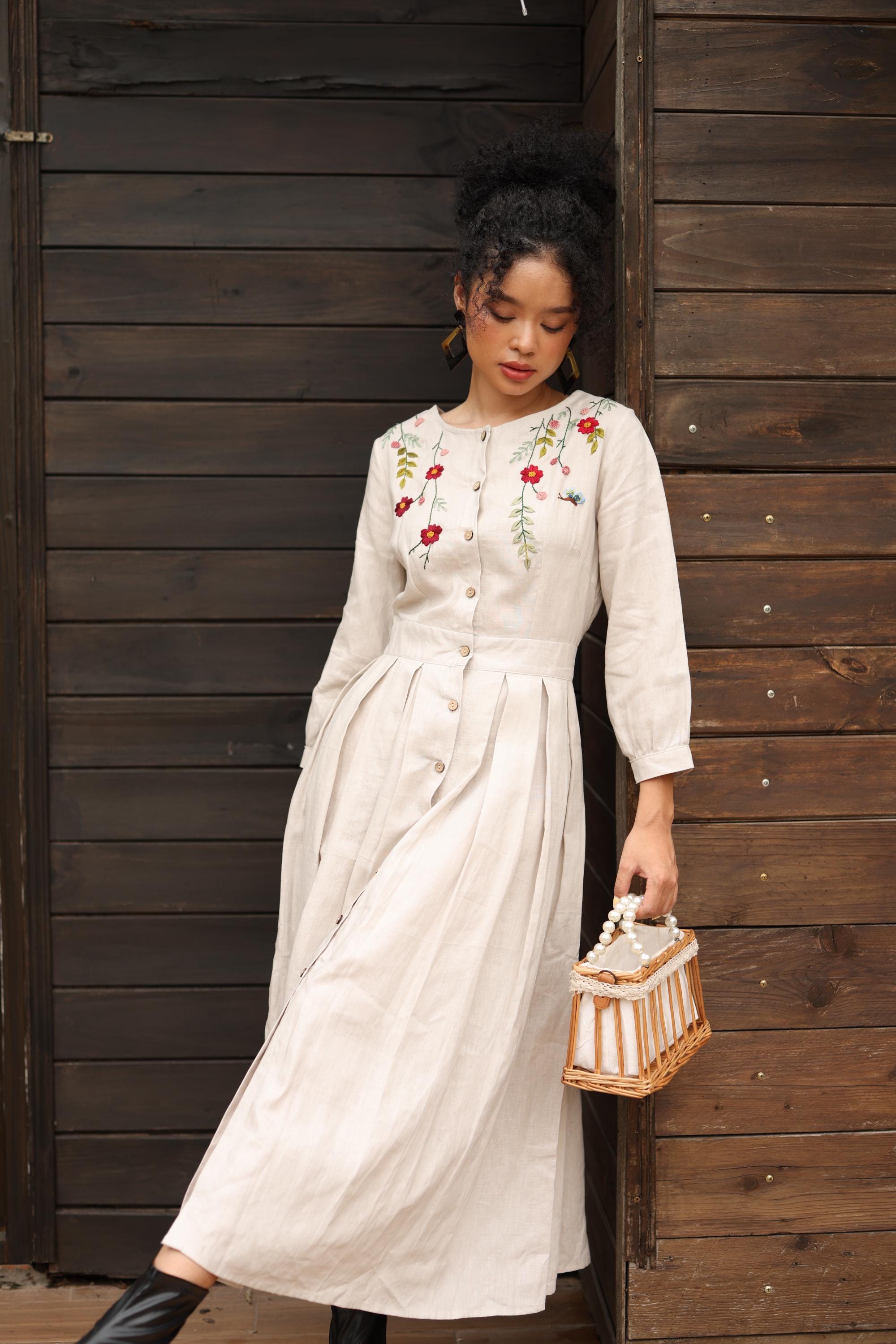 Woman in a dress with floral embroidery standing against a wooden wall.