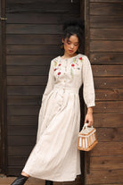 Woman in a dress with floral embroidery standing against a wooden wall.