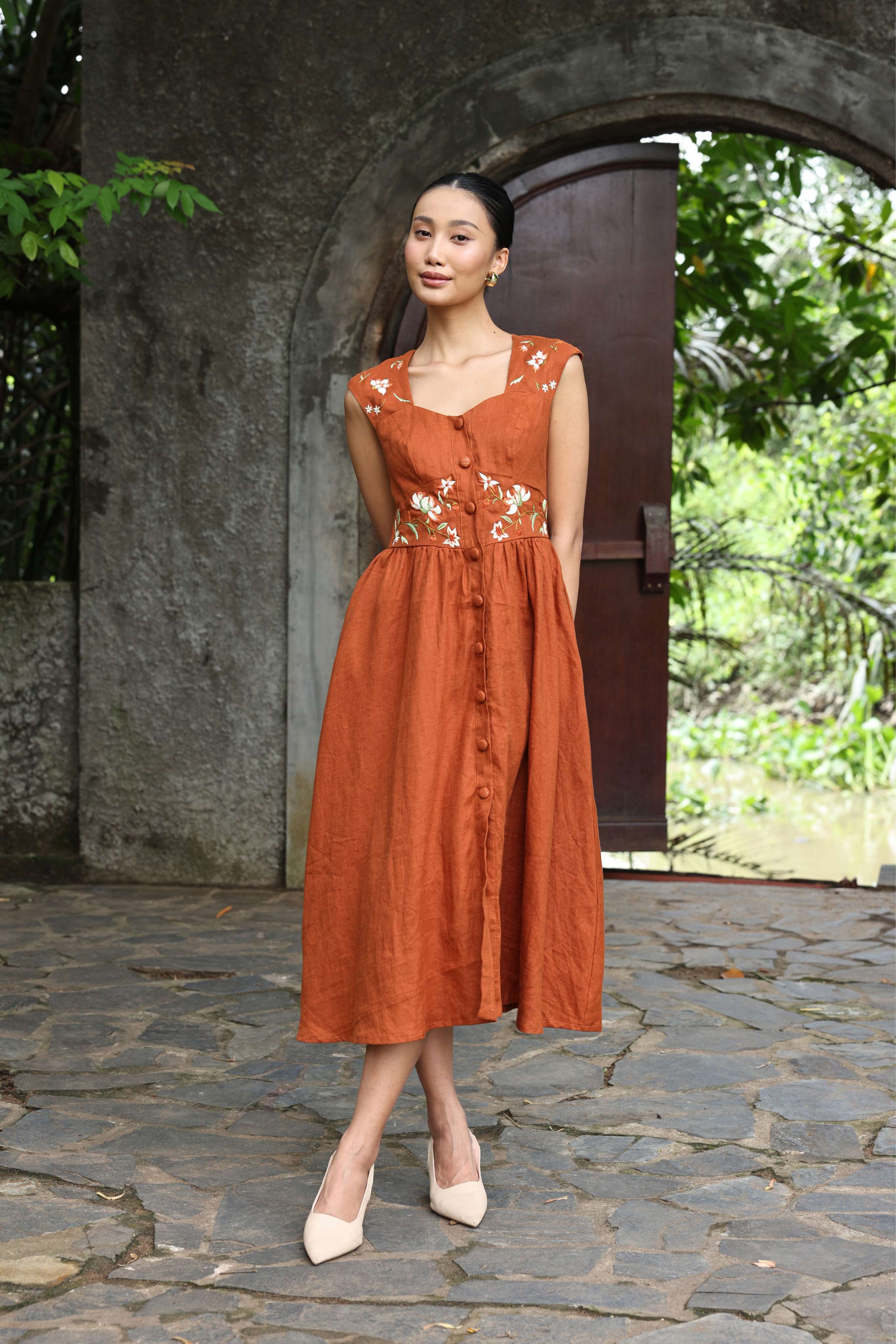 Woman in a rust-colored dress standing outdoors with stone pavement and greenery.