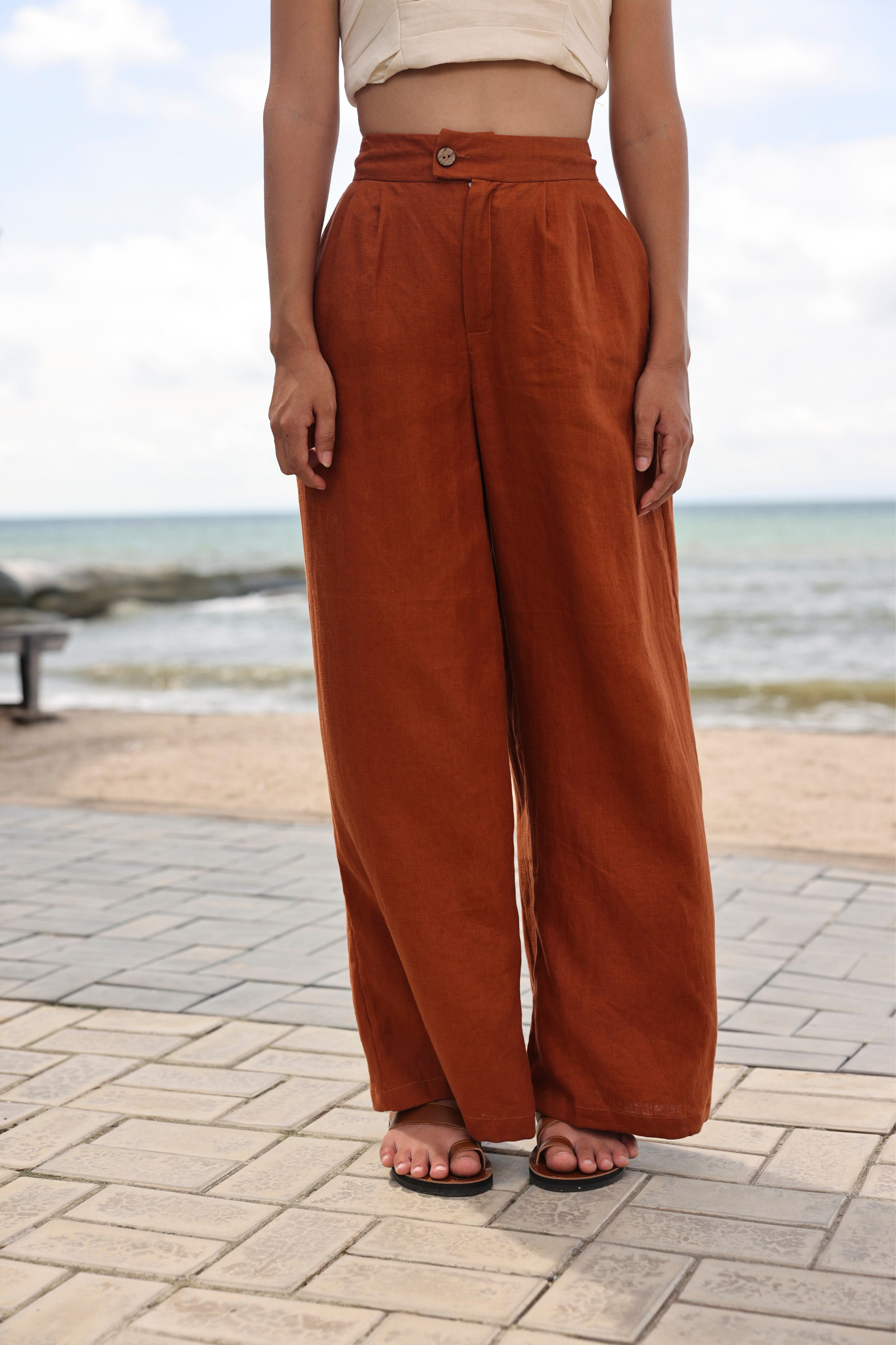 Person wearing rust-colored wide-leg pants standing on a brick pathway with a beach in the background.