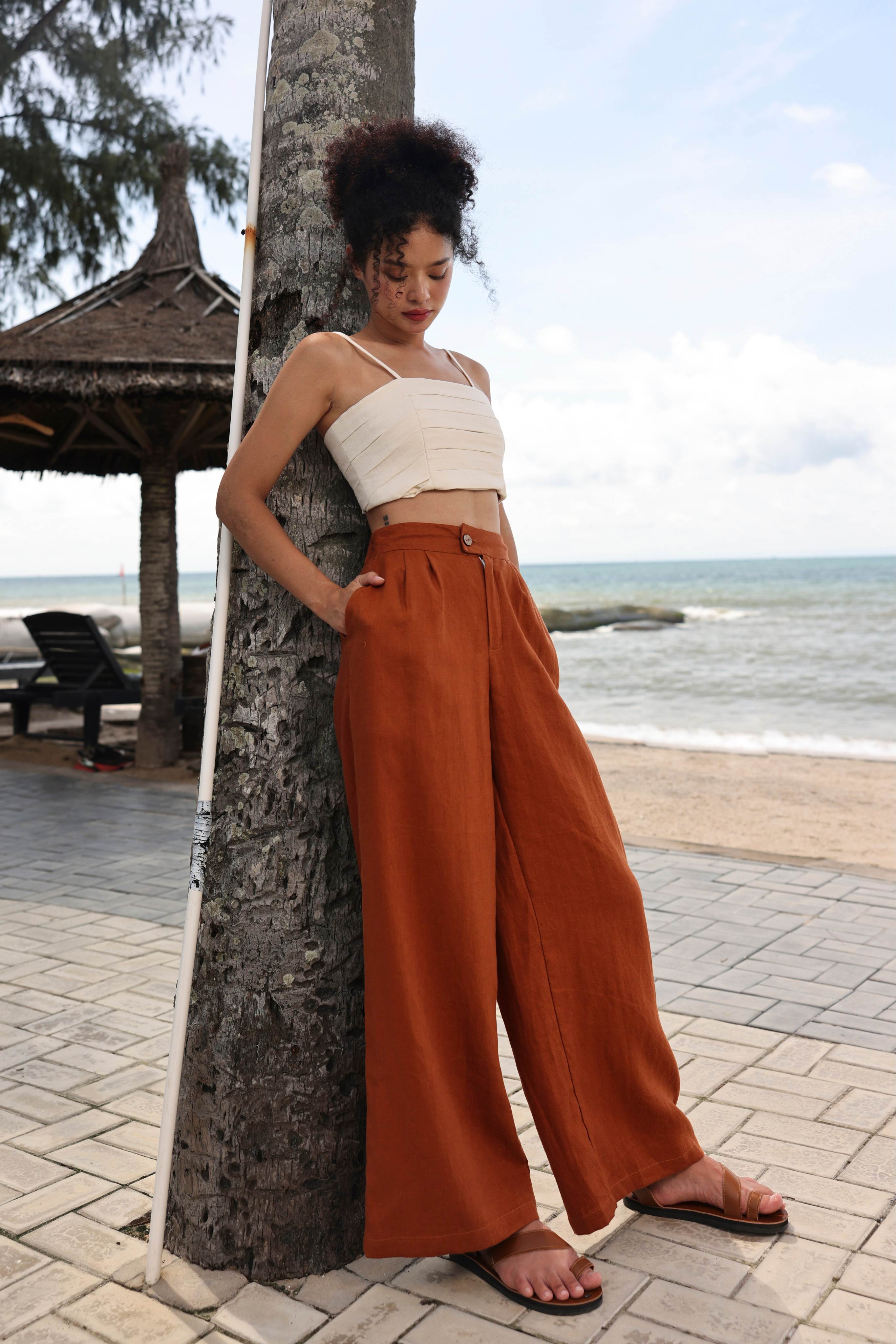 Woman in a white top and orange pants standing by a palm tree on a beach.