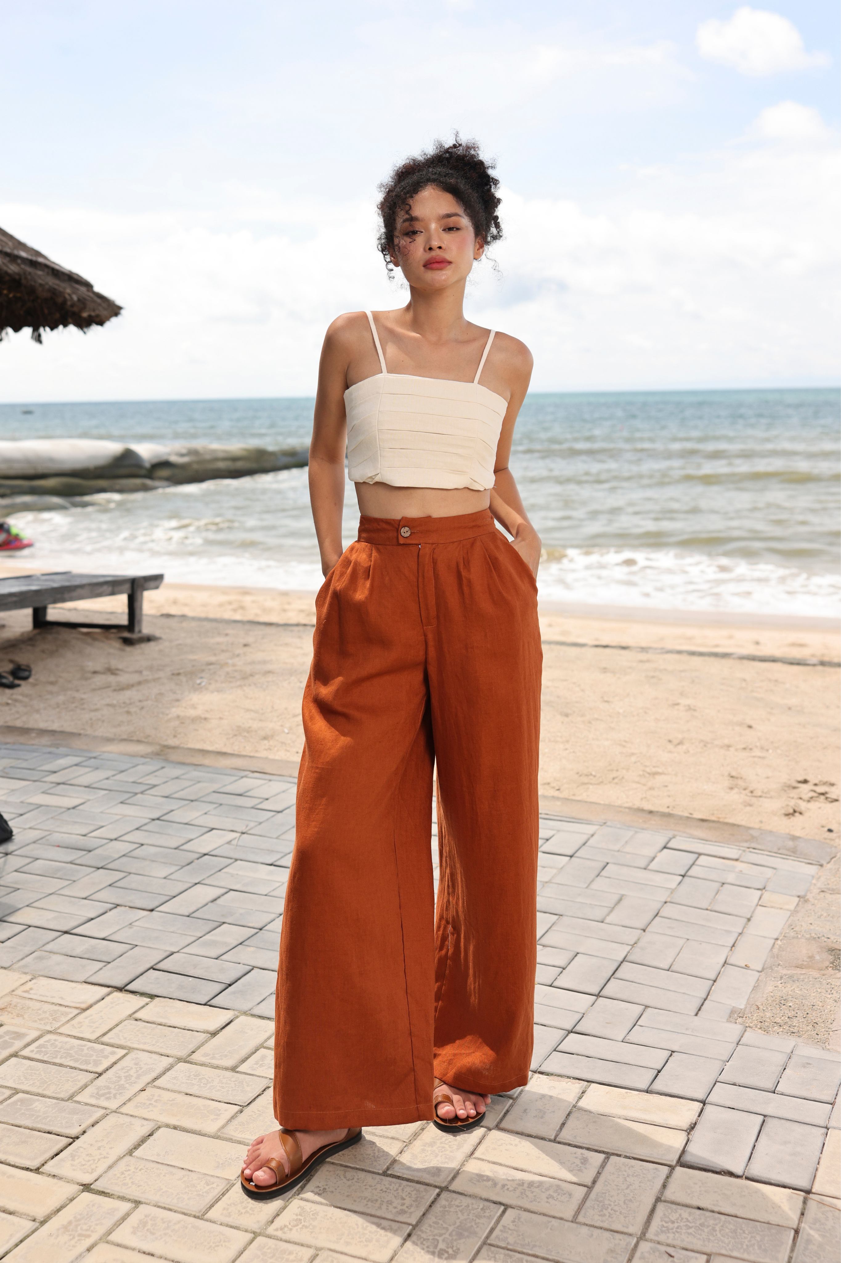 Woman in white top and rust-colored wide-leg pants standing on a beach.