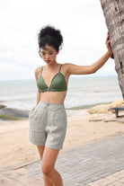 Woman in green bikini top and light gray shorts standing by a tree on a beach.