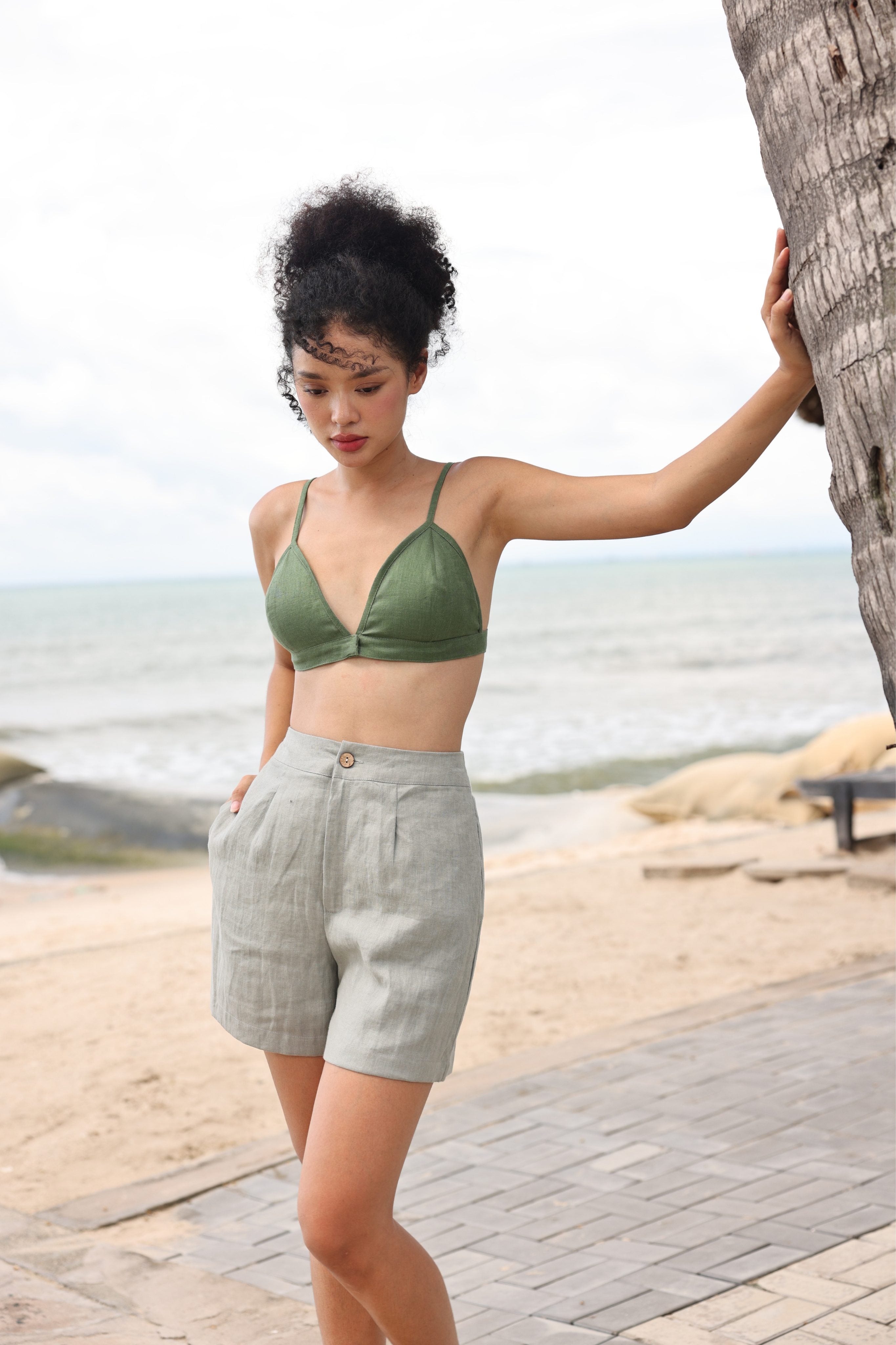Woman in green bikini top and light gray shorts standing by a tree on a beach.