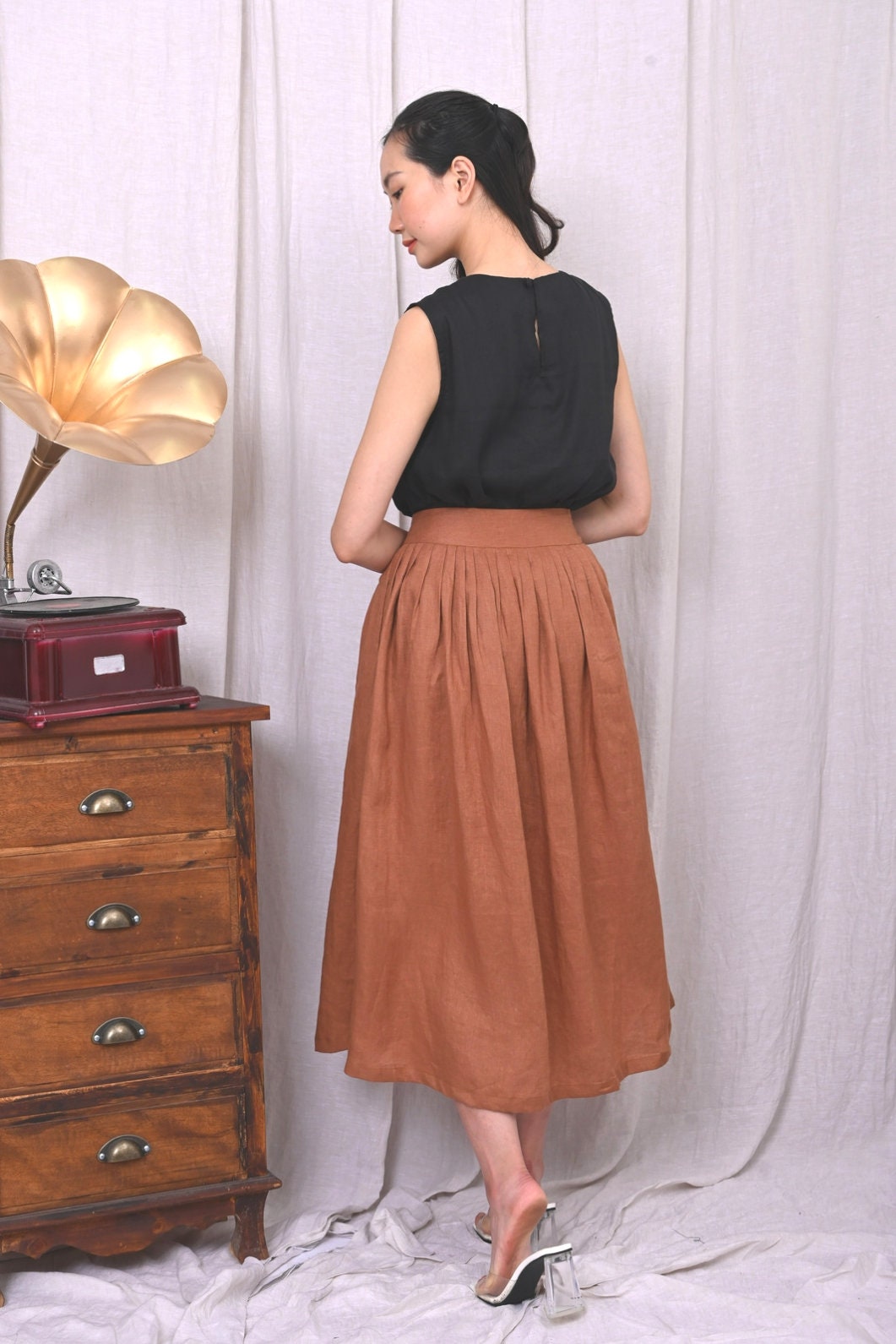 Woman wearing a black top and brown skirt standing next to a wooden cabinet with a lamp.
