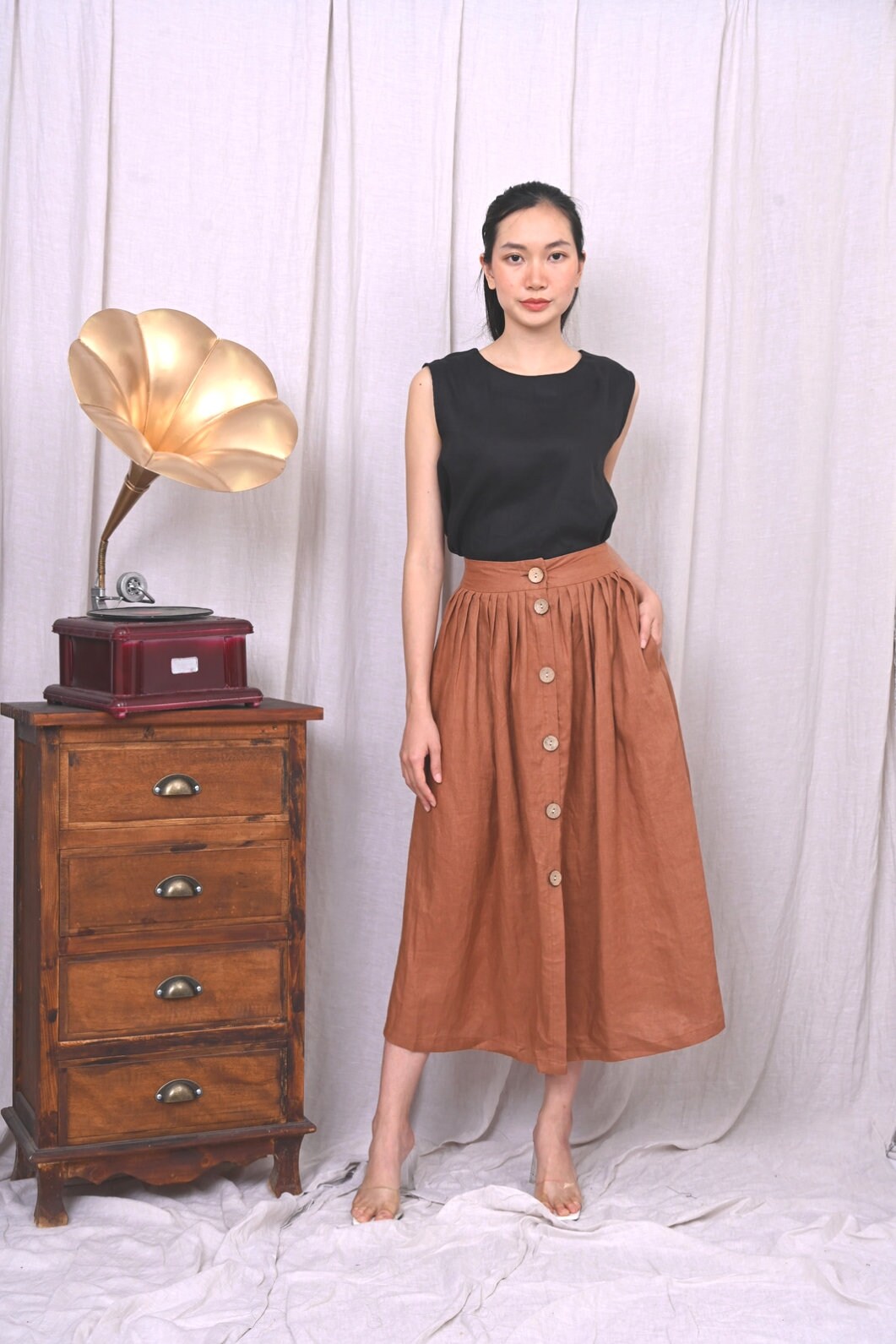 Woman wearing a black top and brown skirt standing next to a vintage wooden record player against a white curtain.