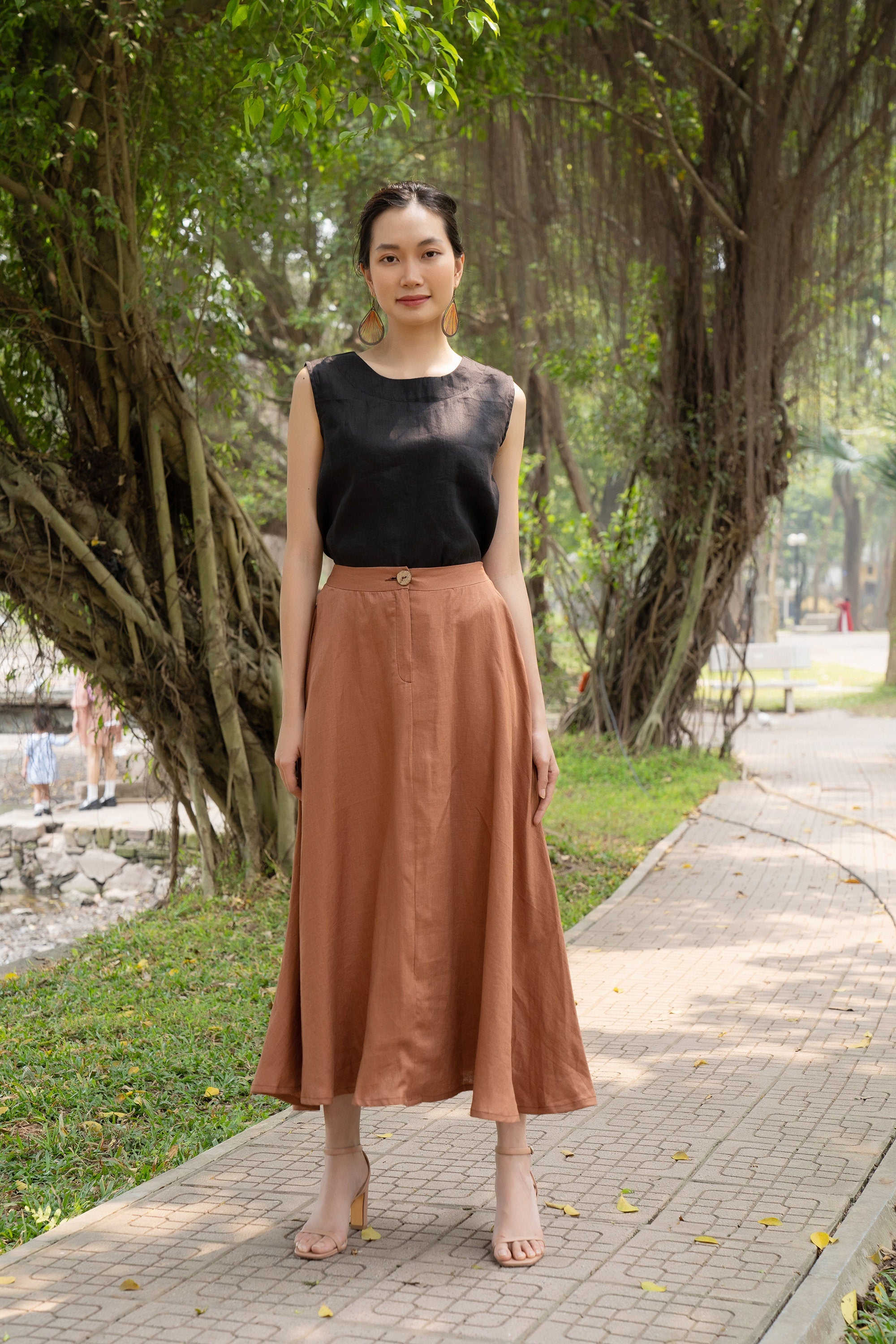 Woman in black top and brown skirt standing on a sidewalk with trees in the background