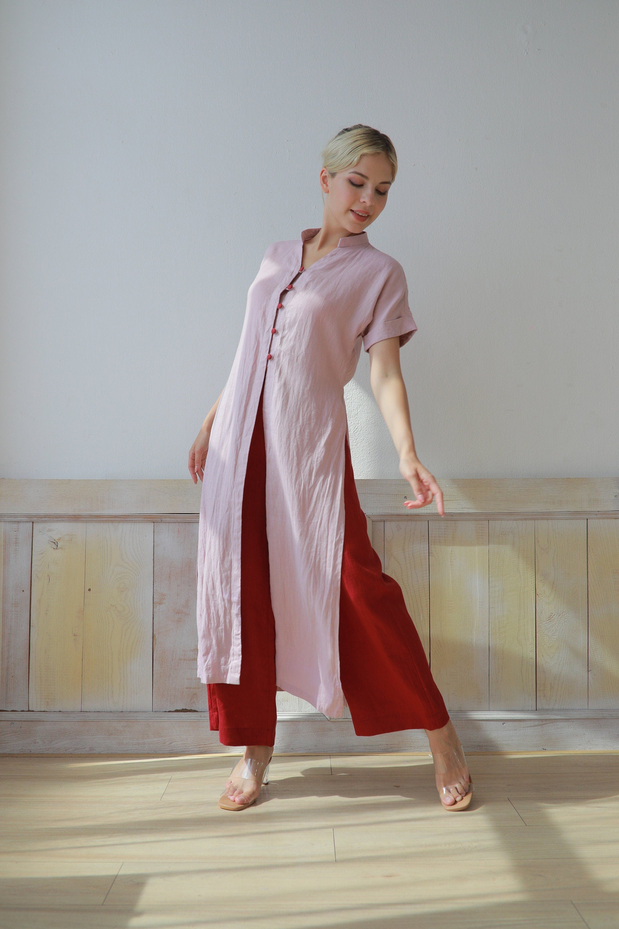 Woman wearing a pink shirt and red pants standing in a room with wooden flooring and a white wall.