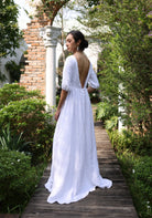 Woman in a white dress with an open back walking on a wooden path in a garden.