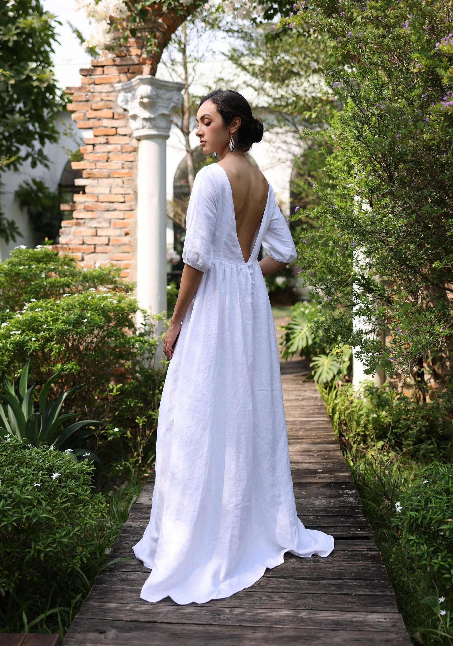 Woman in a white dress with an open back walking on a wooden path in a garden.