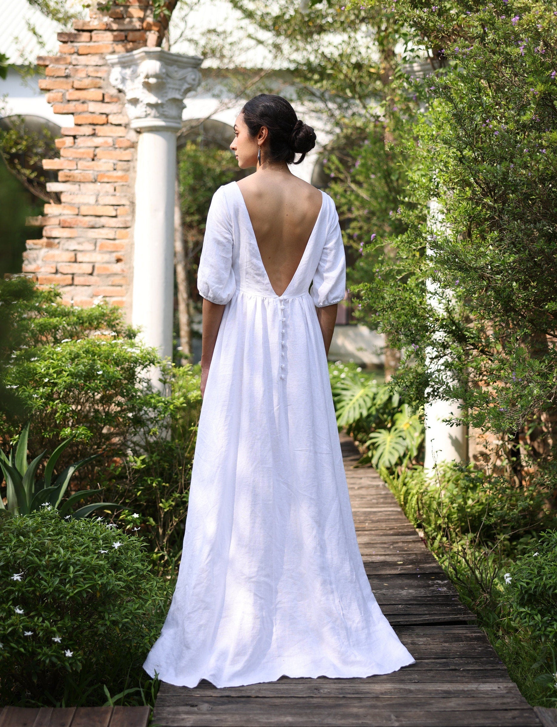 Woman in a white dress standing on a wooden path in a garden.