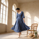 A woman in a blue dress stands in a room with arched windows, her hair pulled back in a bun. A straw hat and a vase of flowers are placed nearby, and a chair is visible in the background.