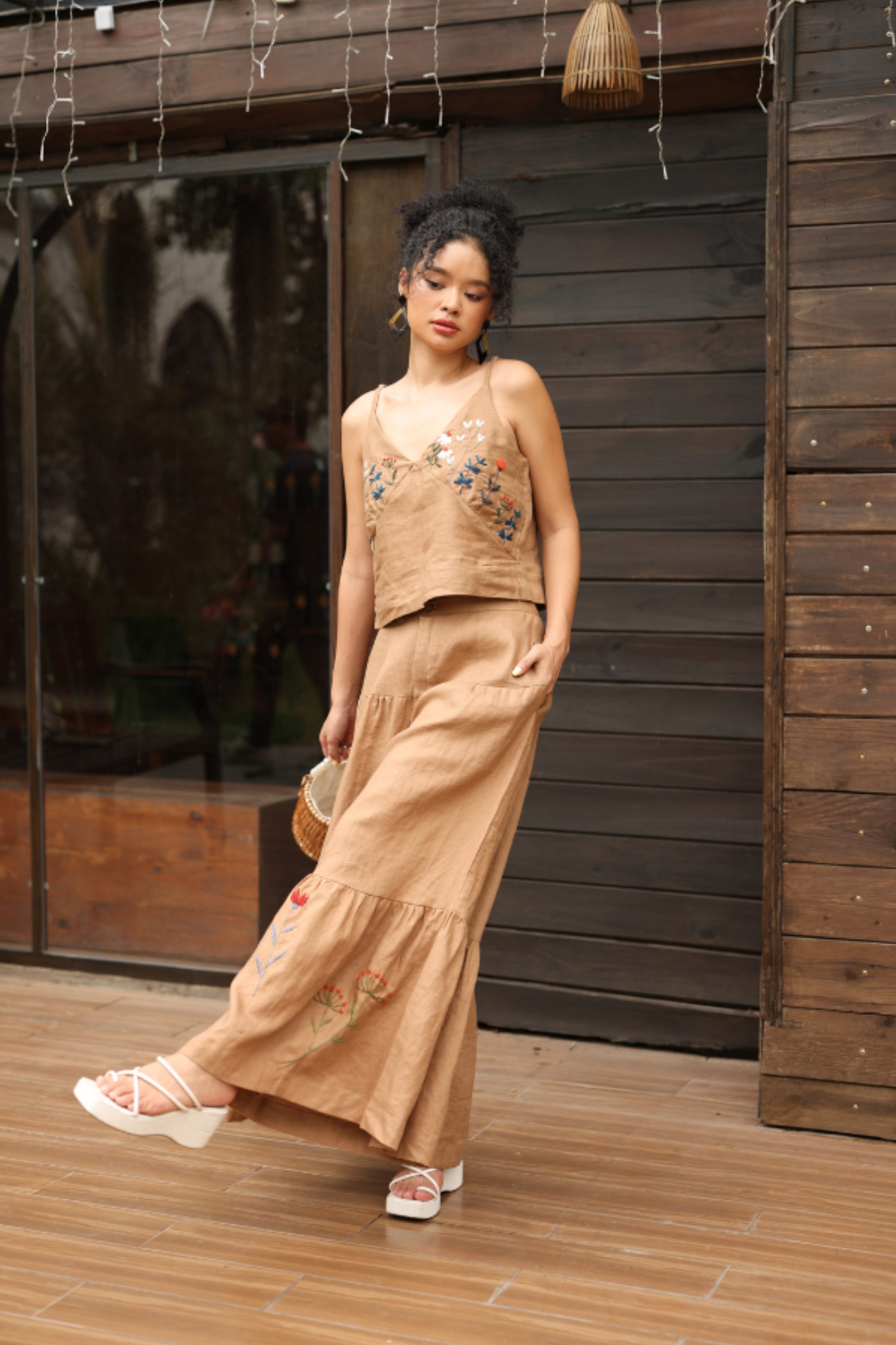 Woman in a beige outfit with floral details standing in front of a wooden wall.