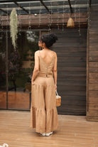 Woman in a beige crop top and pants standing on a wooden deck with a wooden wall and plants in the background.