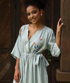 Woman wearing a light blue dress with floral embroidery standing against a wooden background