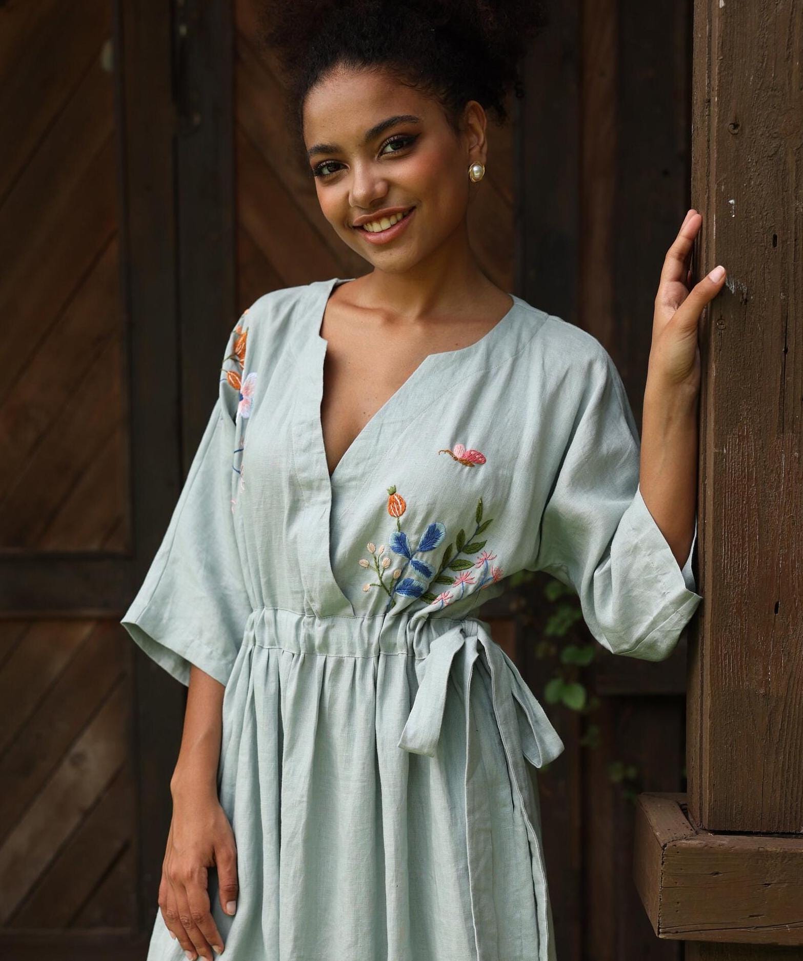 Woman wearing a light blue dress with floral embroidery standing against a wooden background