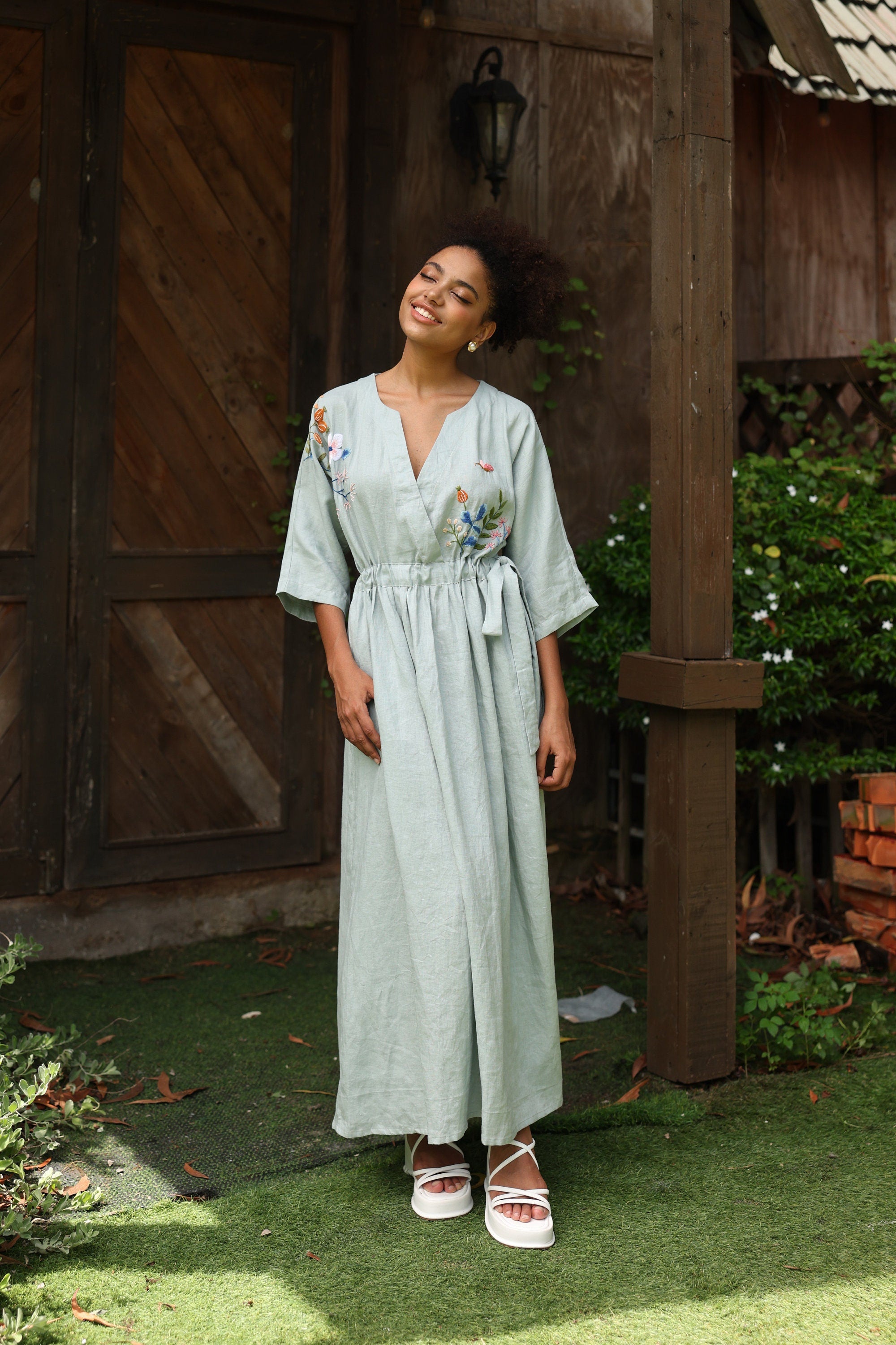 Woman in a light green dress standing outdoors near a wooden building.