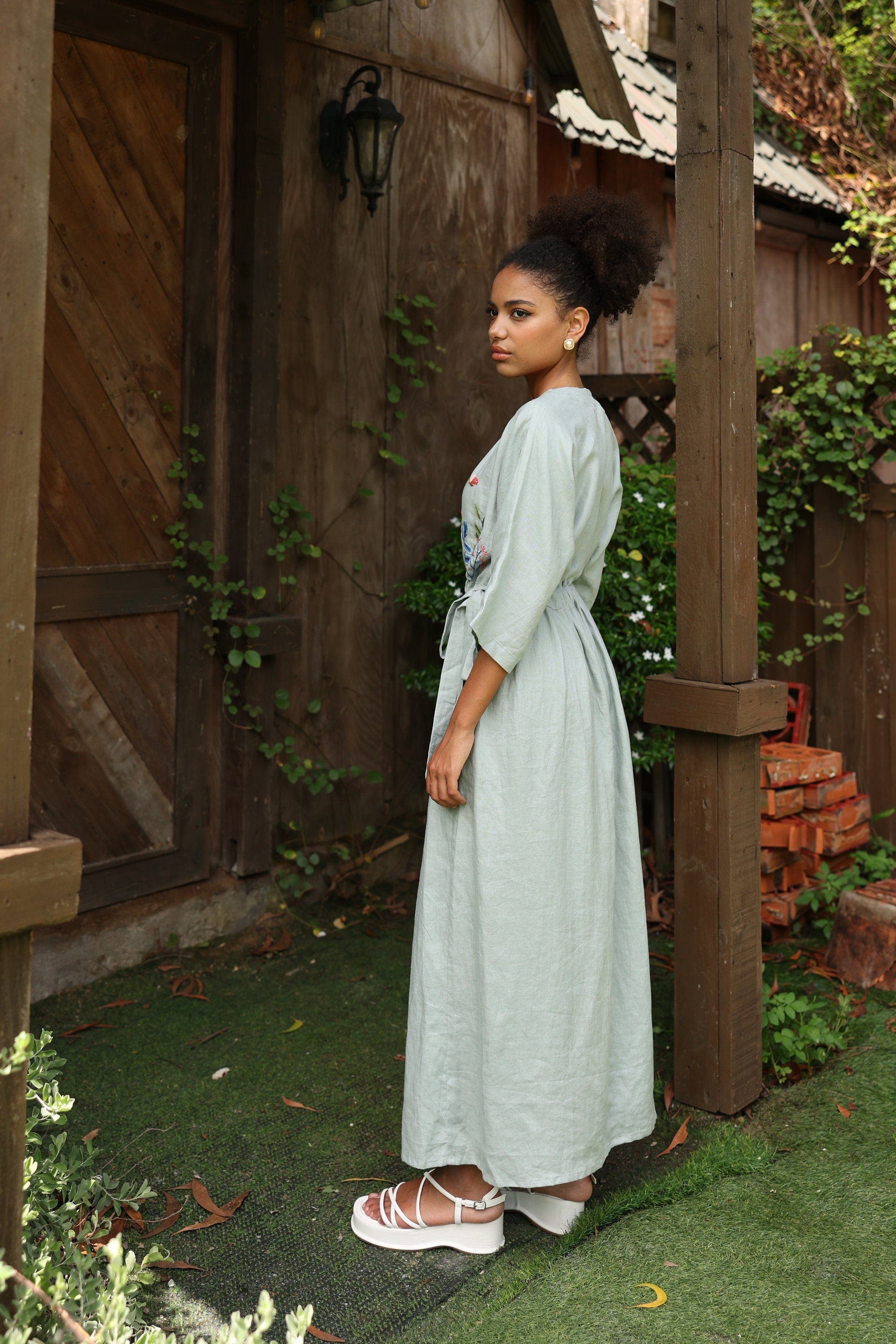 Woman in a dress standing in front of a wooden building with plants around.