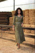 Woman in a green dress with floral embroidery standing in a barn with hay bales.