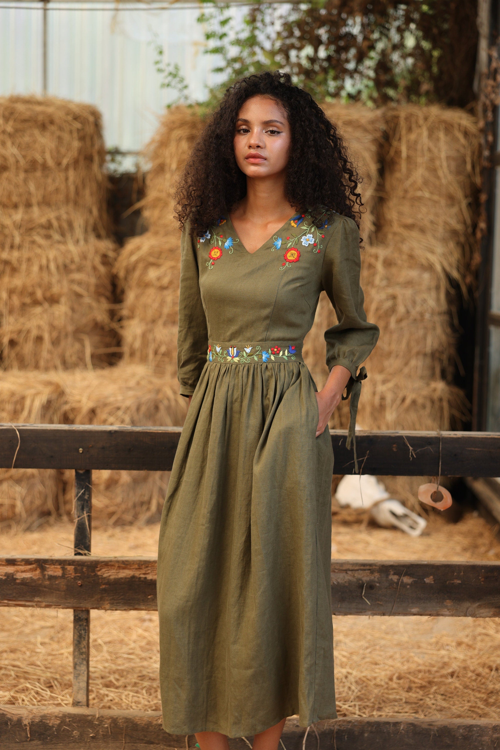 Woman in a green dress with floral embellishments standing in front of hay bales.