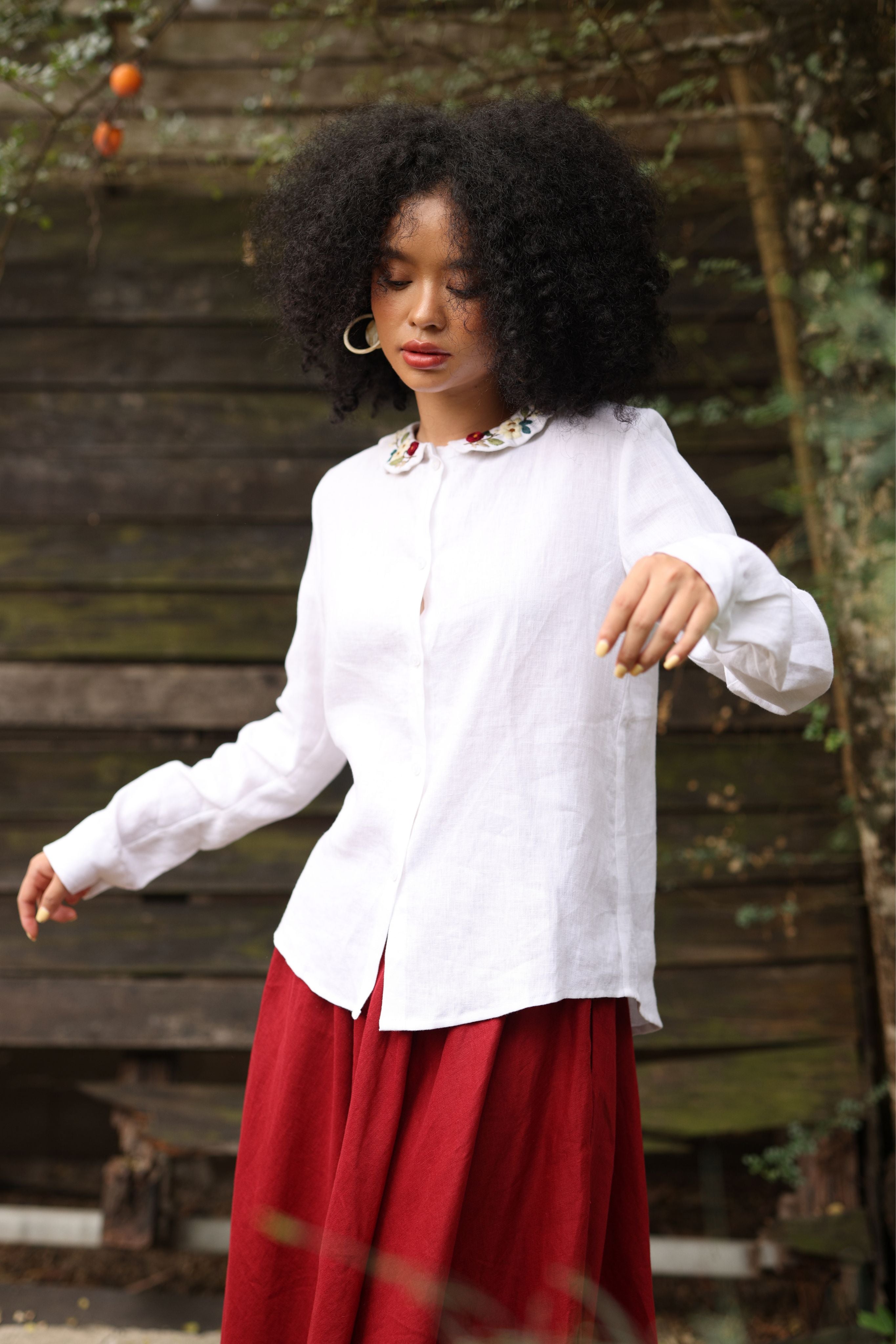 Woman wearing a white blouse and red skirt standing outdoors with a wooden fence and plants in the background.